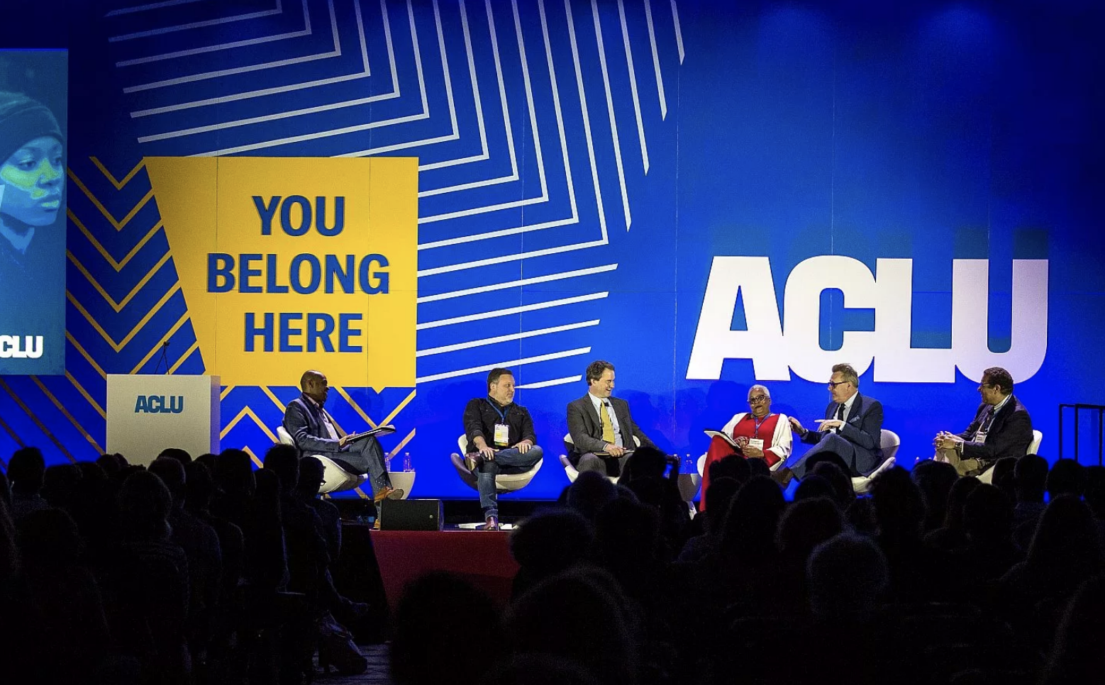 A panel of six people engage in a discussion on a stage with the ACLU logo prominently displayed in the background. A large yellow sign reads "YOU BELONG HERE." An audience is partially visible in the foreground. The backdrop features a blue and white geometric design.