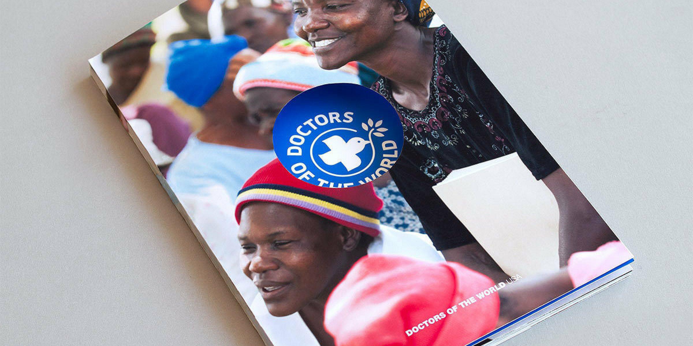 A brochure titled "Doctors of the World" lies on a grey surface. The cover features smiling individuals wearing colorful clothing and headscarves. The prominently visible logo consists of a white cross inside a blue circle with the organization's name.