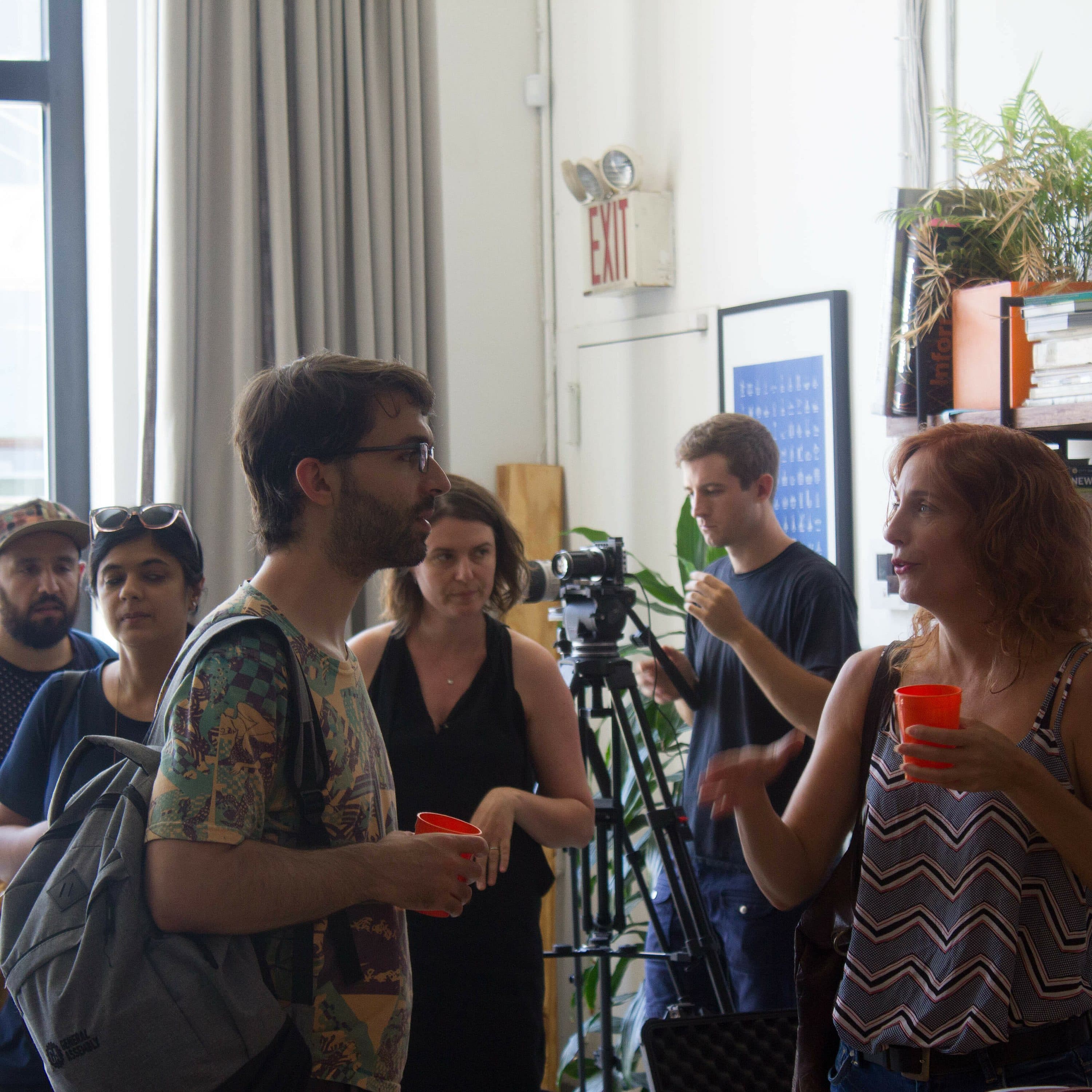 A group of seven adults in casual attire are gathered indoors, engaging in conversation. One man with a backpack and glasses talks to a woman holding a red cup. Behind them, a person operates a camera, and a bookshelf filled with books is visible on the right.