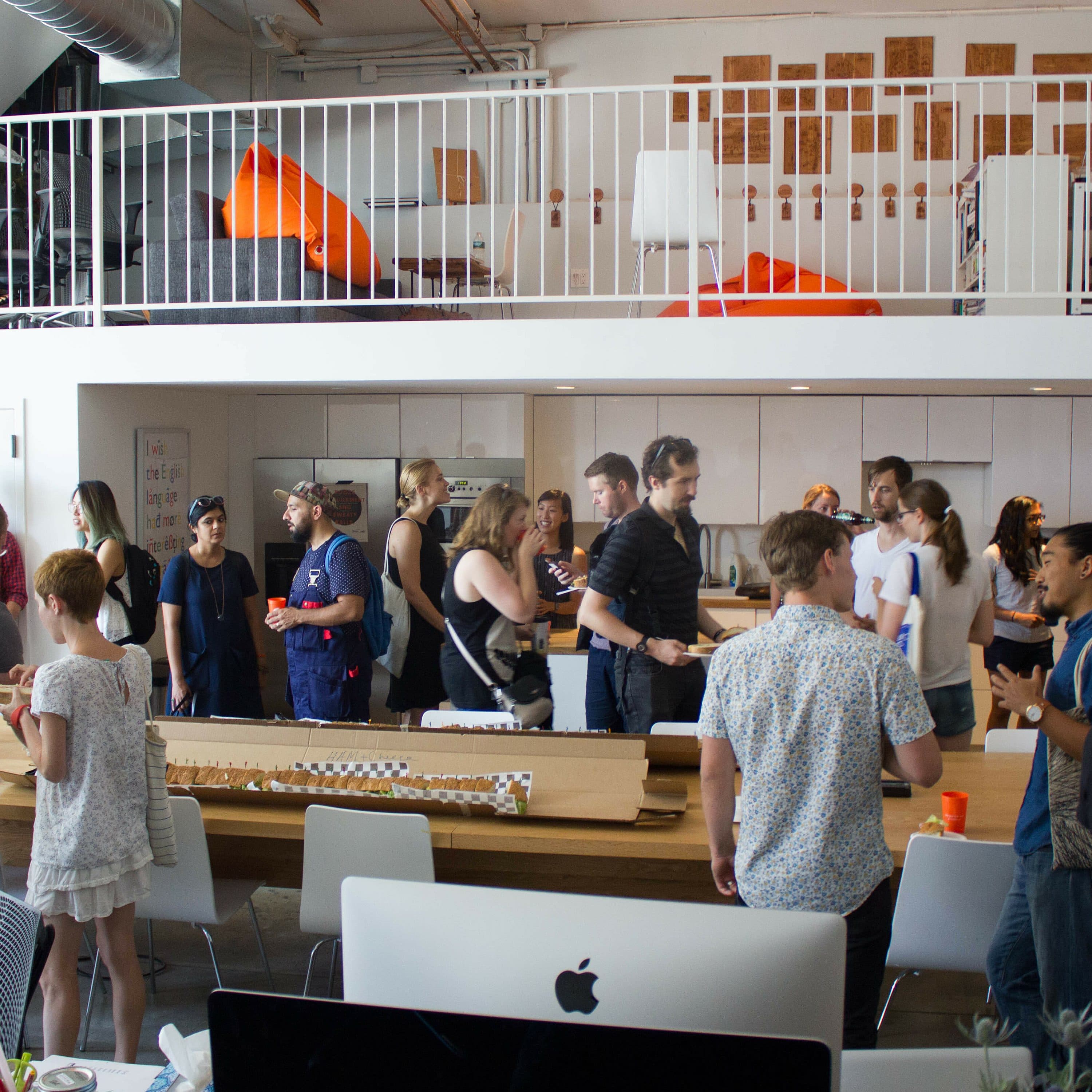 A diverse group of people socializing at a casual indoor gathering. Some are standing, engaging in conversations, while others are seated or walking around. The space is modern and open, with a second-floor loft visible above and various pieces of furniture and decor.