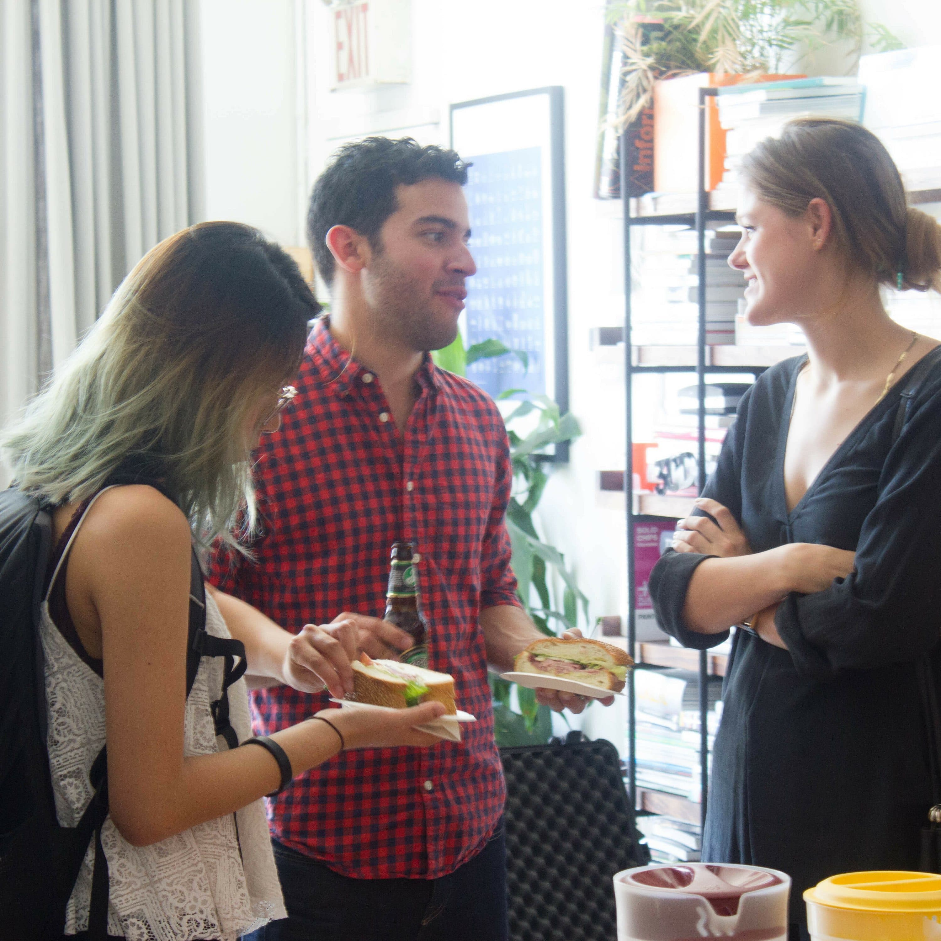 A group of people in a casual setting, conversing and eating. One person holds two sandwich containers and another holds a beverage. Shelves filled with books and plants are in the background. The atmosphere appears friendly and relaxed.