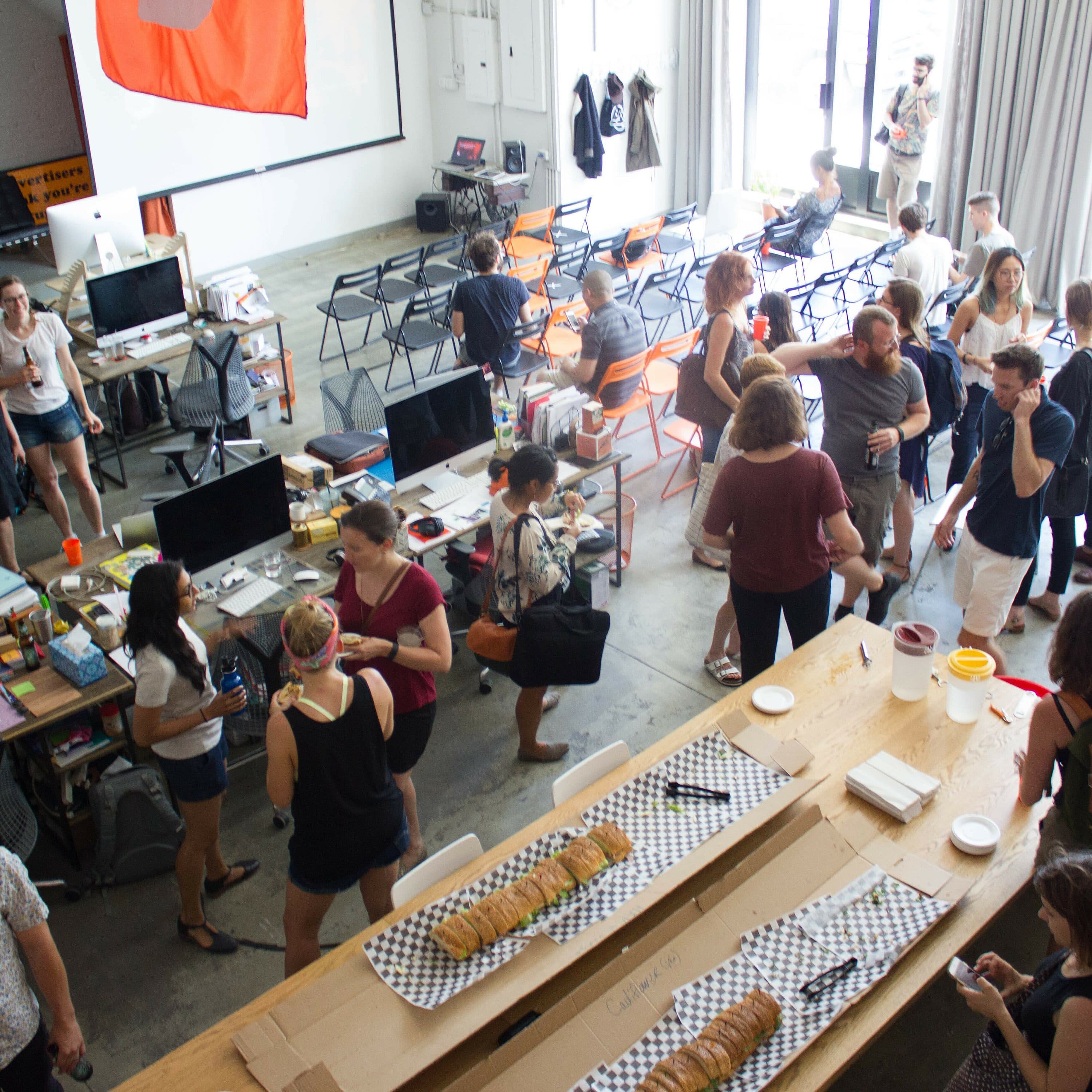 A large, well-lit room filled with people socializing. The room has long tables with food, several computers, and other office equipment. Some people are standing and talking, while others sit at tables. There is a large orange banner on the wall.