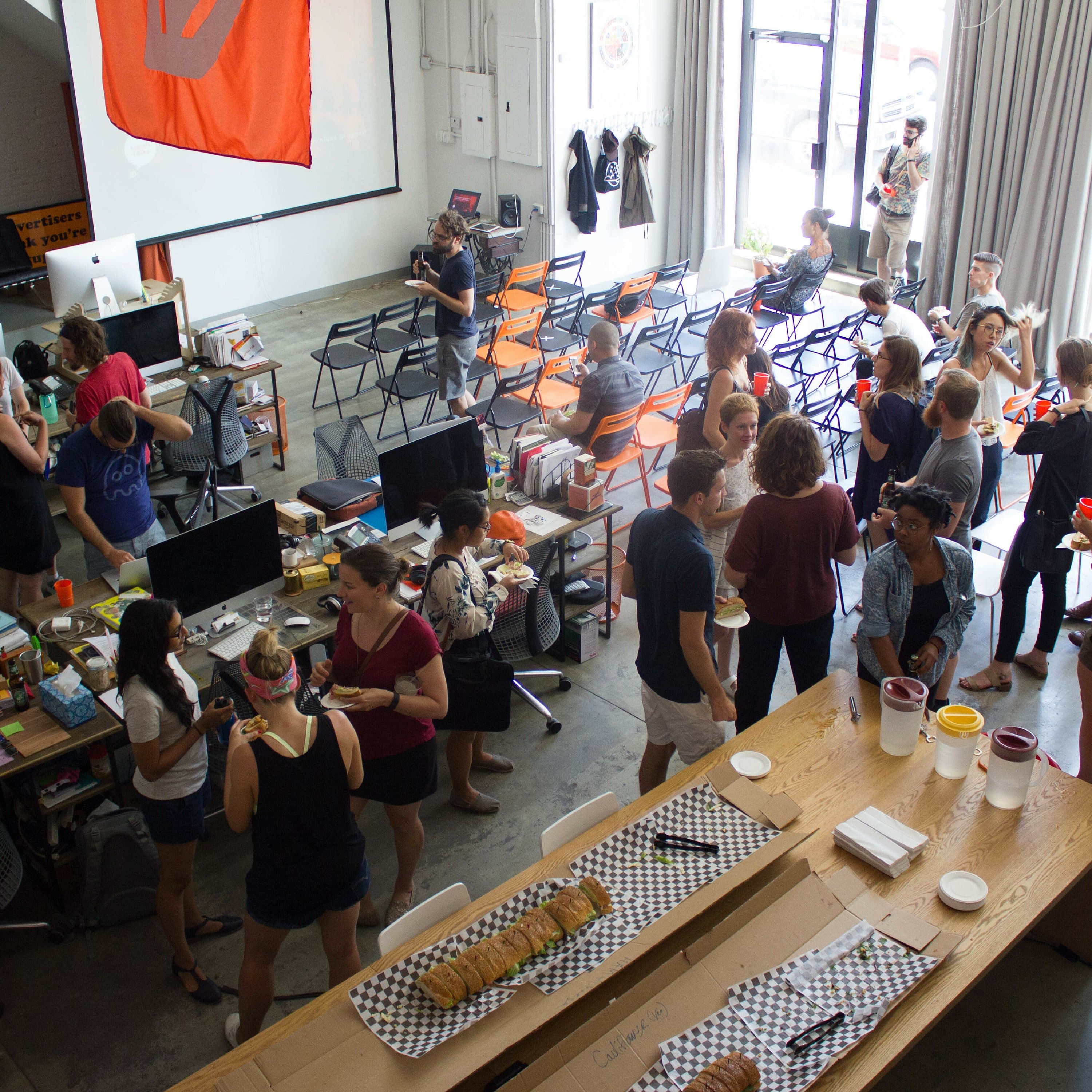 A large group of people mingles inside a spacious, modern office with high ceilings and large windows. Tables are set with food, and various items such as computers and decorations are visible. There are chairs arranged for seating, and a relaxed atmosphere is evident.