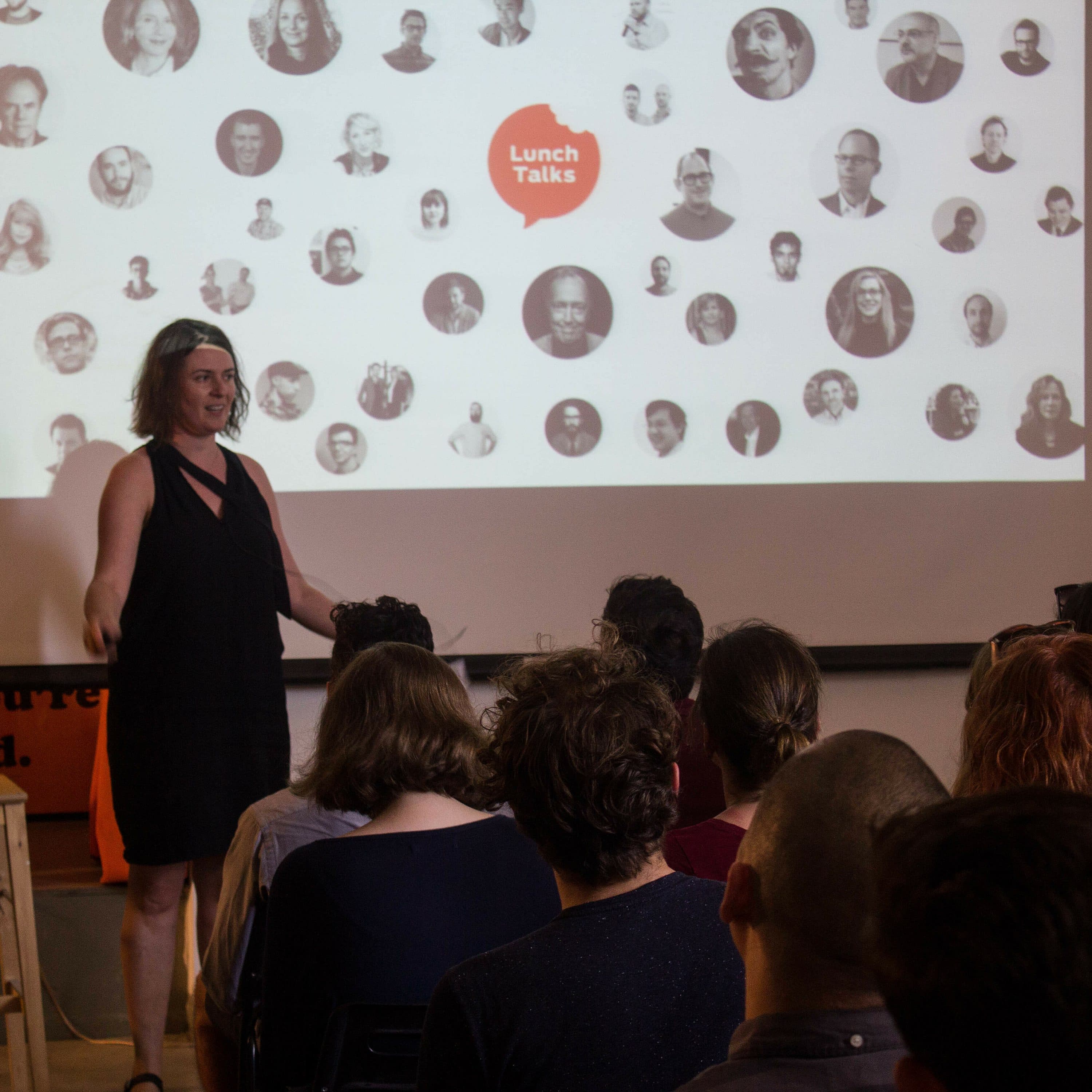 A woman in a black dress stands in front of a seated audience, presenting at an event. Behind her is a screen displaying a collage of black-and-white headshots with the heading "Lunch Talks" in the center. The venue has a casual, informal atmosphere.