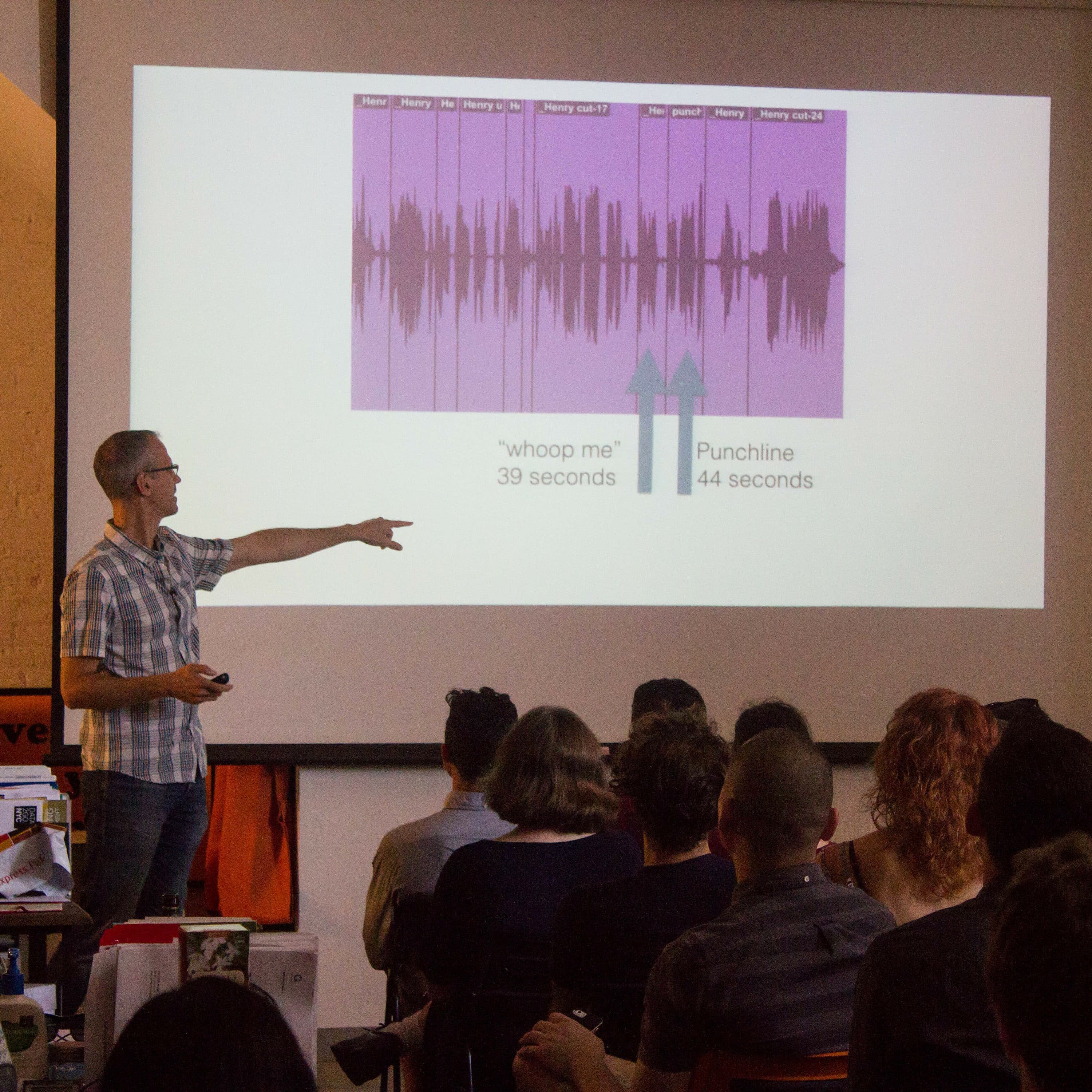 A person wearing glasses and a checked shirt stands at the front of a classroom, pointing at a projected slide. The slide shows a graph with sections labeled "whoop me?" and "Punchline," marked with time durations of 39 seconds and 44 seconds respectively. An audience watches intently.