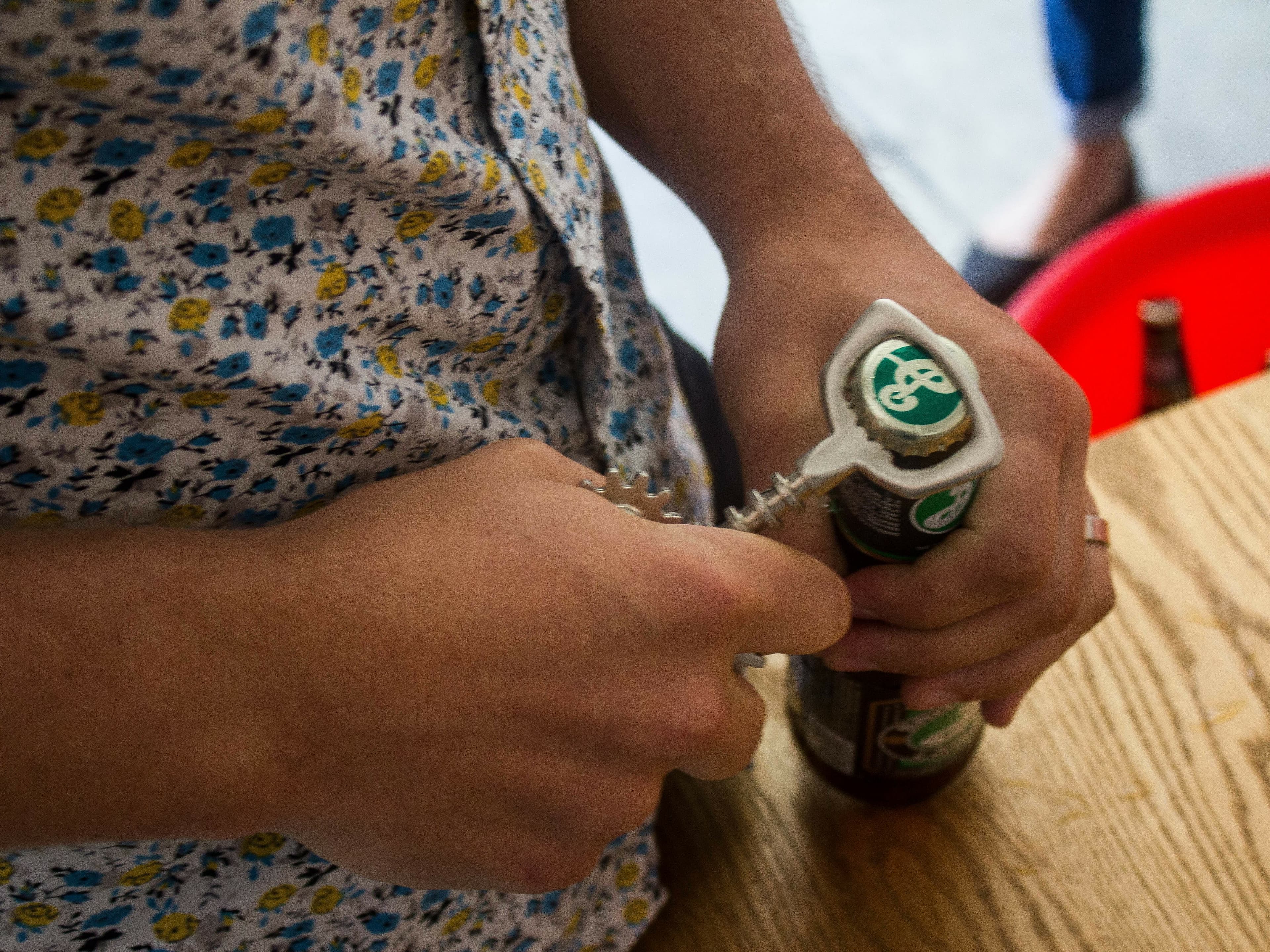 A person wearing a floral shirt uses a corkscrew to open a bottle of Brooklyn Lager beer on a wooden table. The background is slightly blurred, showing part of another person and a chair.