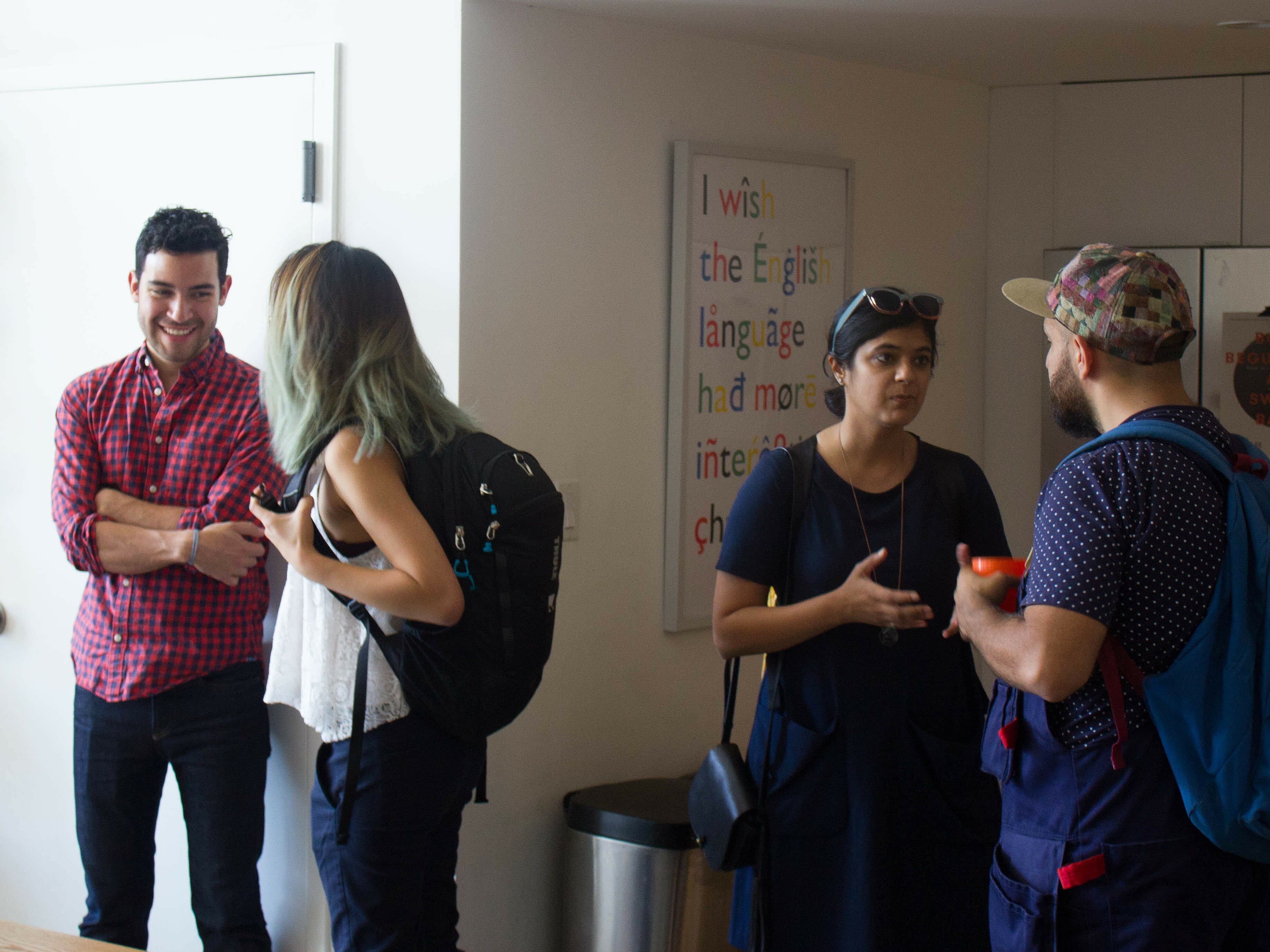 Five people are gathered in a casual indoor setting. Three are standing and talking, while one is looking at their phone. Another person is entering the room, smiling. A colorful sign in the background reads: "I wish the English language had more interesting choices.