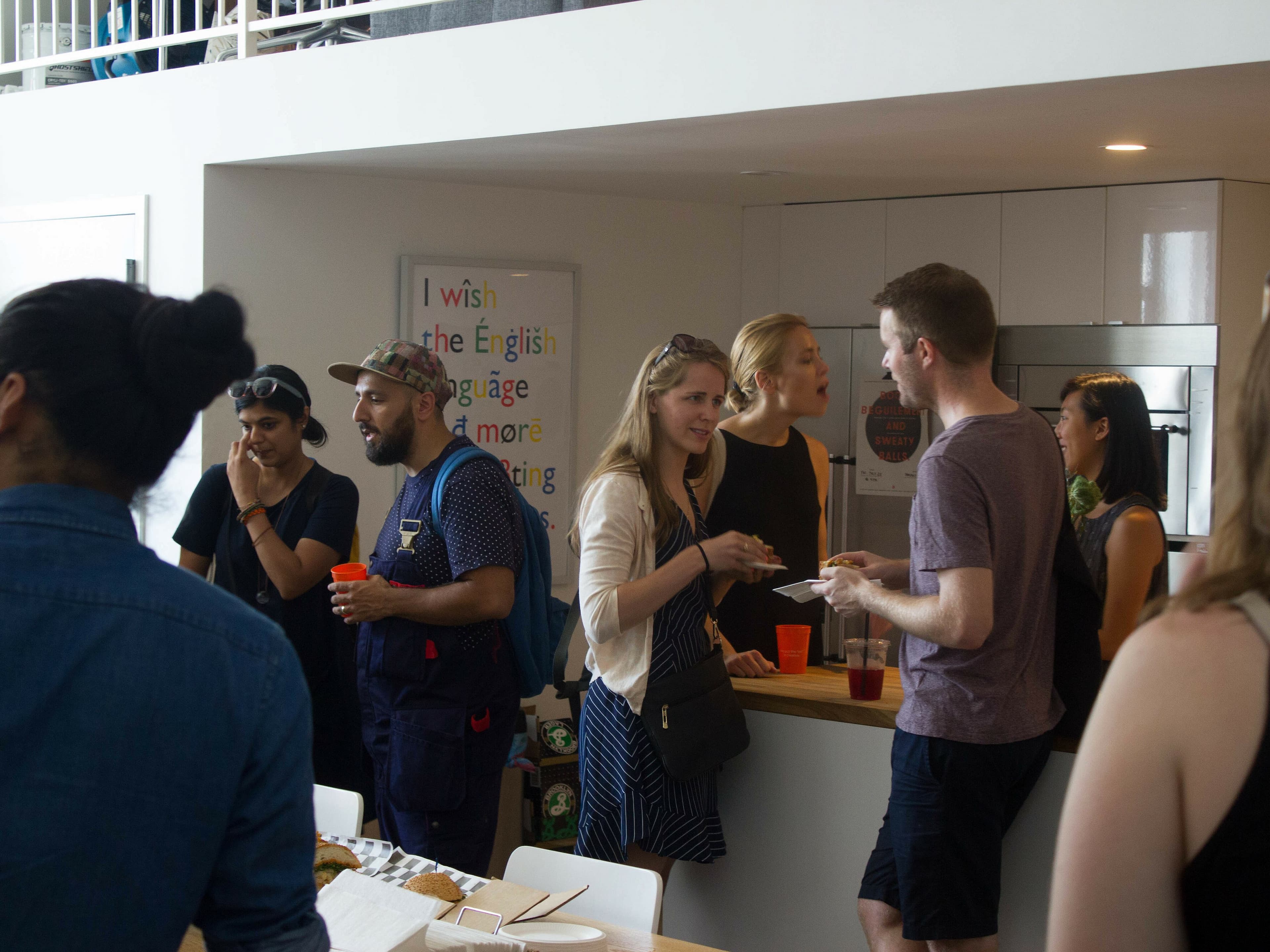 A group of people socializing at a gathering in a modern kitchen space. Some are holding drinks and plates of food. A poster in the background reads, "I wish the English language had more exciting." The atmosphere appears casual and lively.