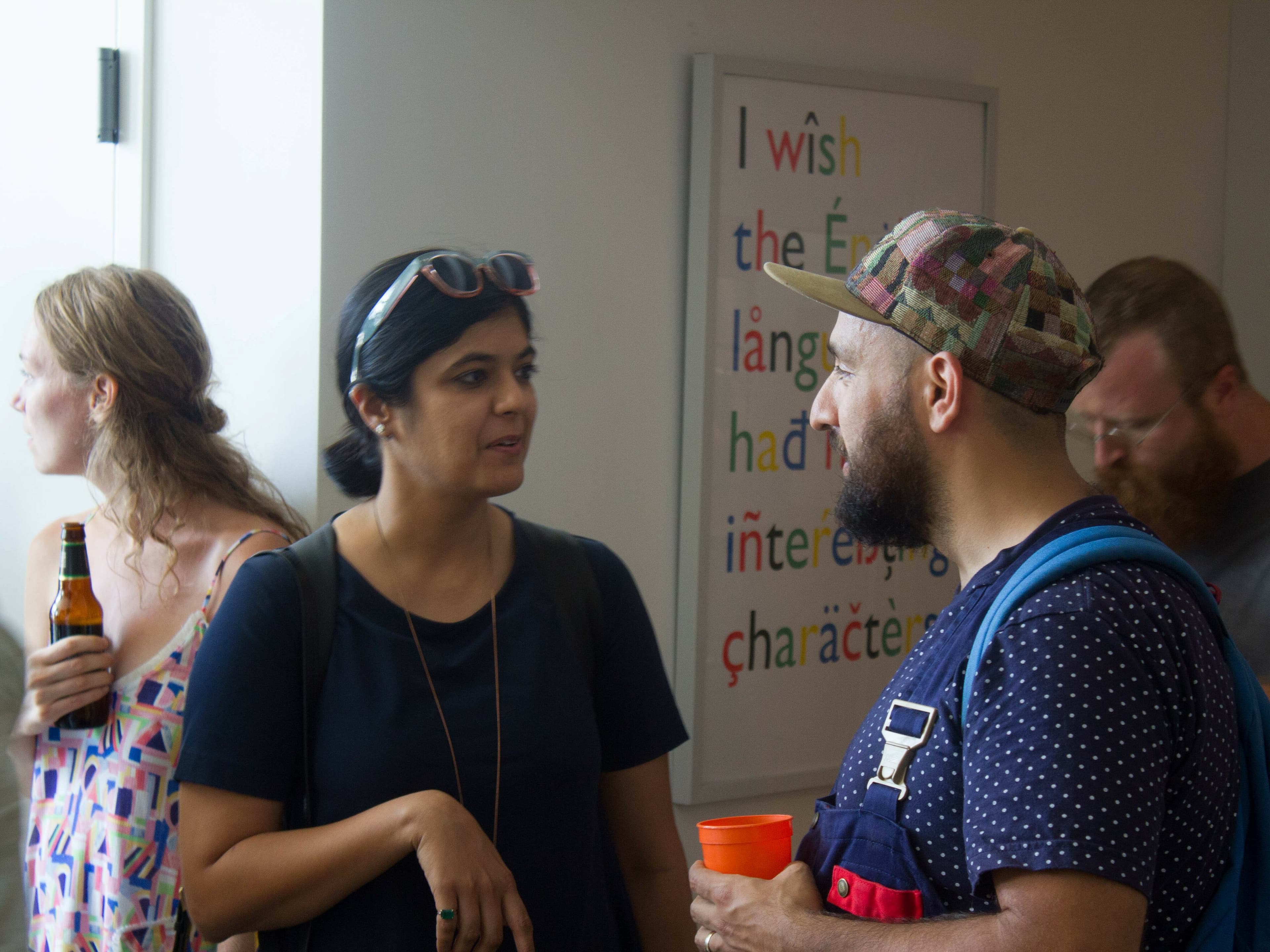 A group of people is gathered in a room having conversations. In the foreground, a woman with sunglasses on her head and a man wearing a patterned cap and holding an orange cup are talking. A colorful poster with abstract text is on the wall behind them.