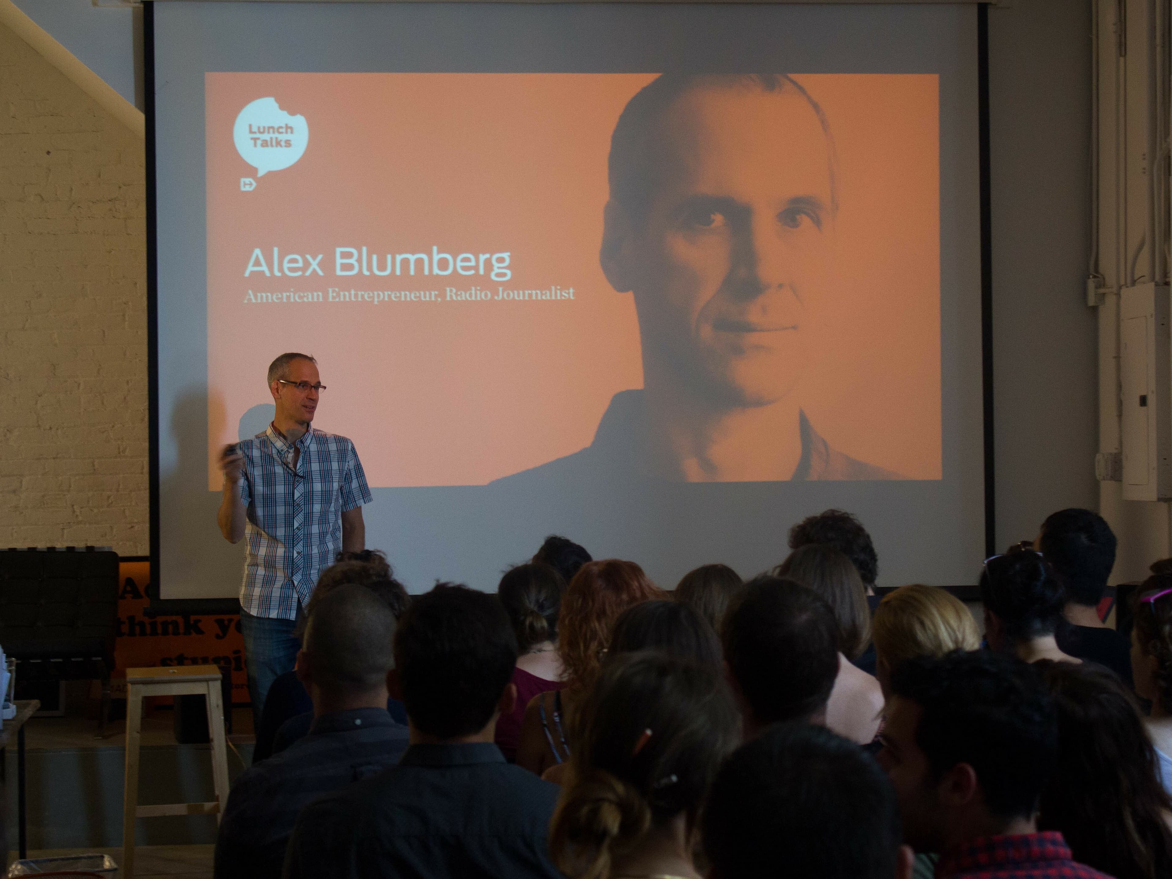 A speaker is presenting in front of a seated audience. Behind him is a large screen displaying a photo and name, "Alex Blumberg," along with subtitles, "American Entrepreneur, Radio Journalist" and "Lunch Talks." The venue is a well-lit room.