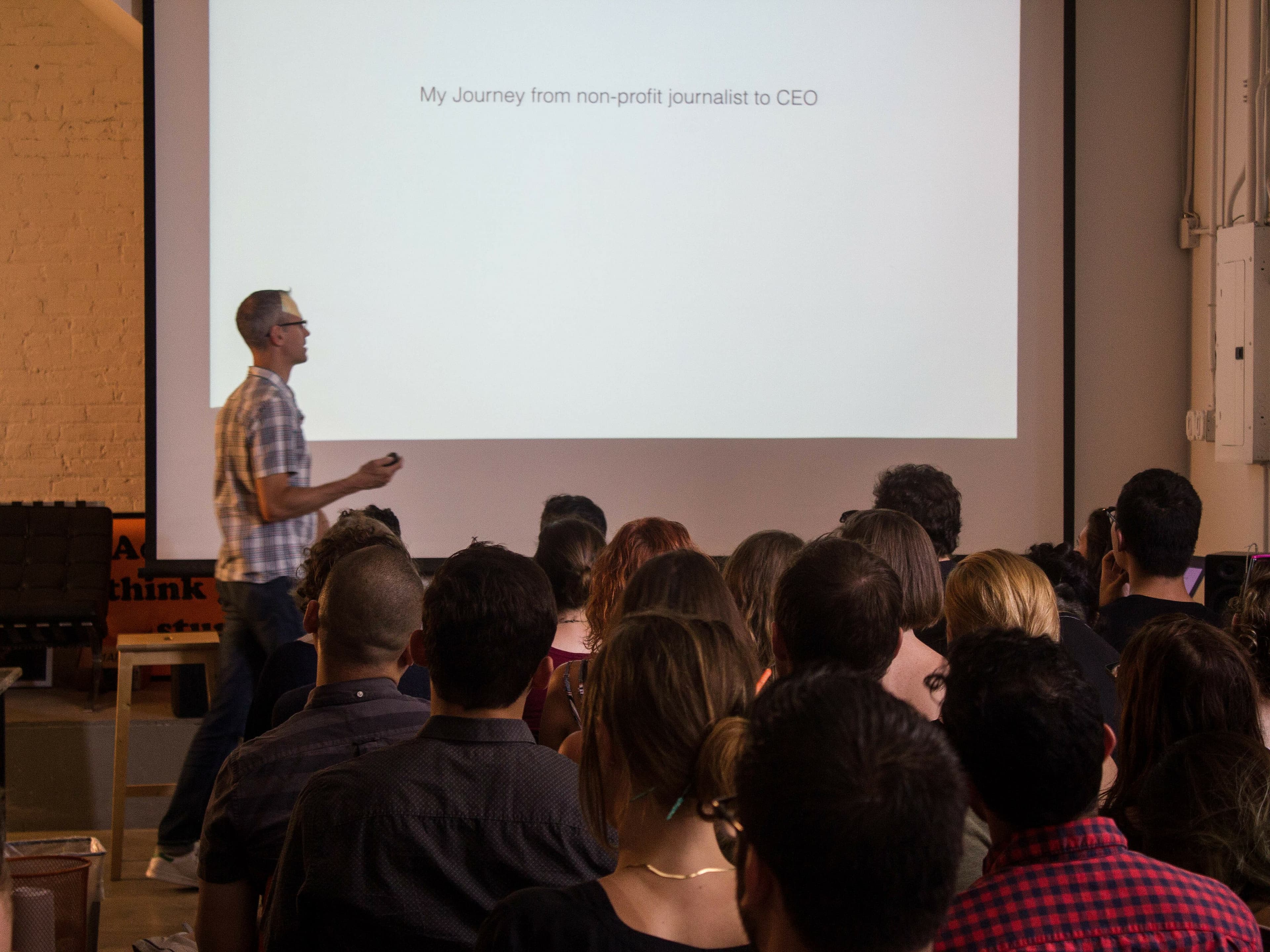 A man in a plaid shirt is presenting to a seated audience in a room with a large screen displaying the slide title "My Journey from non-profit journalist to CEO." People in the audience are facing the presenter, and the room has a casual, modern setup.