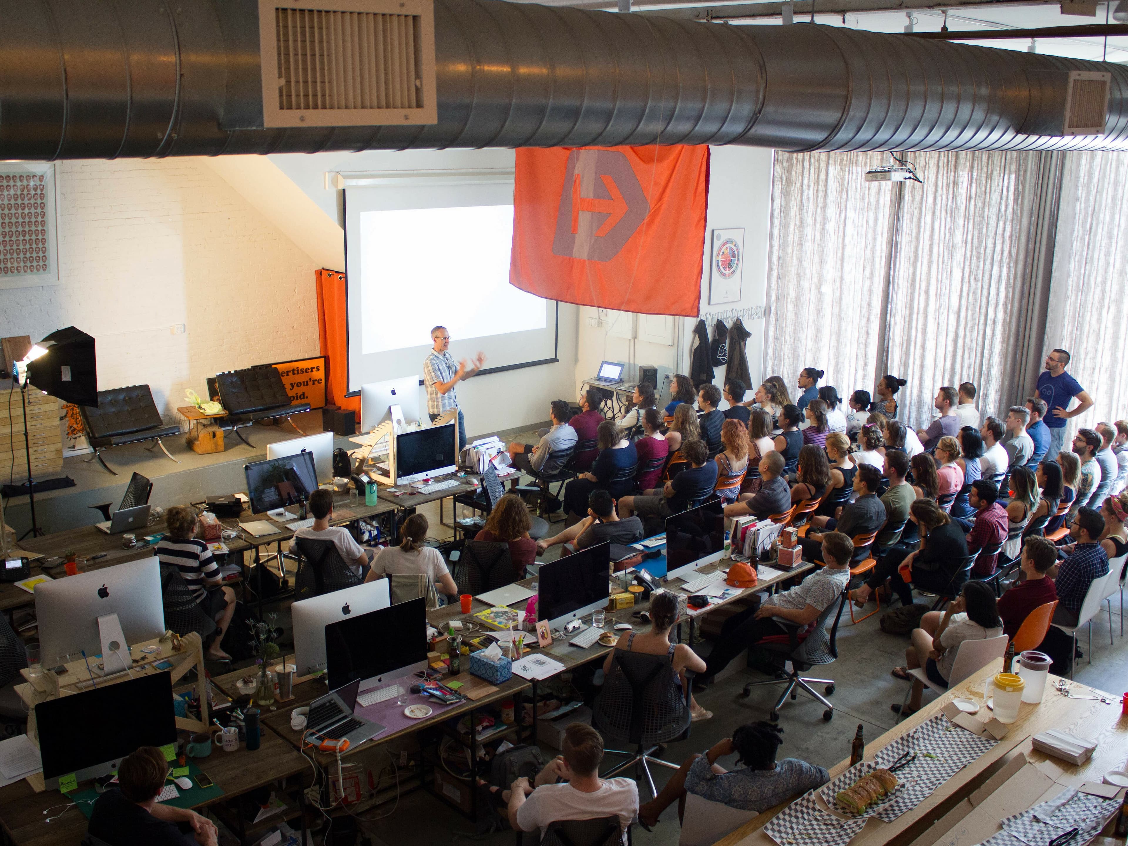 A group of people seated and attentively listening to a speaker in a brightly lit, industrial-style office space. A large screen displays a presentation at the front, and various desks with computers and equipment surround the audience. An orange flag hangs on the wall.