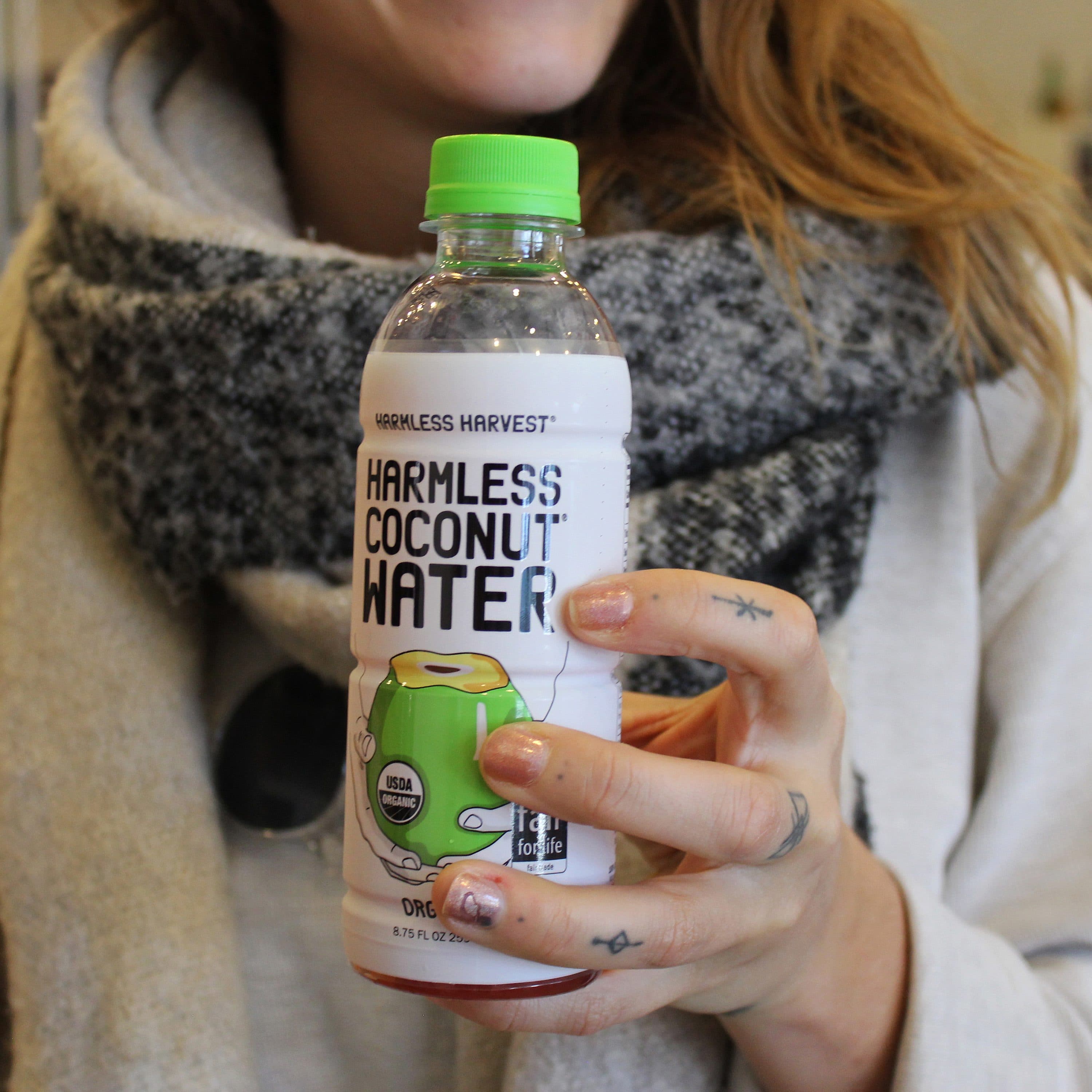 A person with light brown hair holds a bottle of Harmless Harvest Coconut Water. The person is wearing a grey scarf and a light-colored top. The focus is on the bottle, which has a green label with a coconut illustration and the brand name.