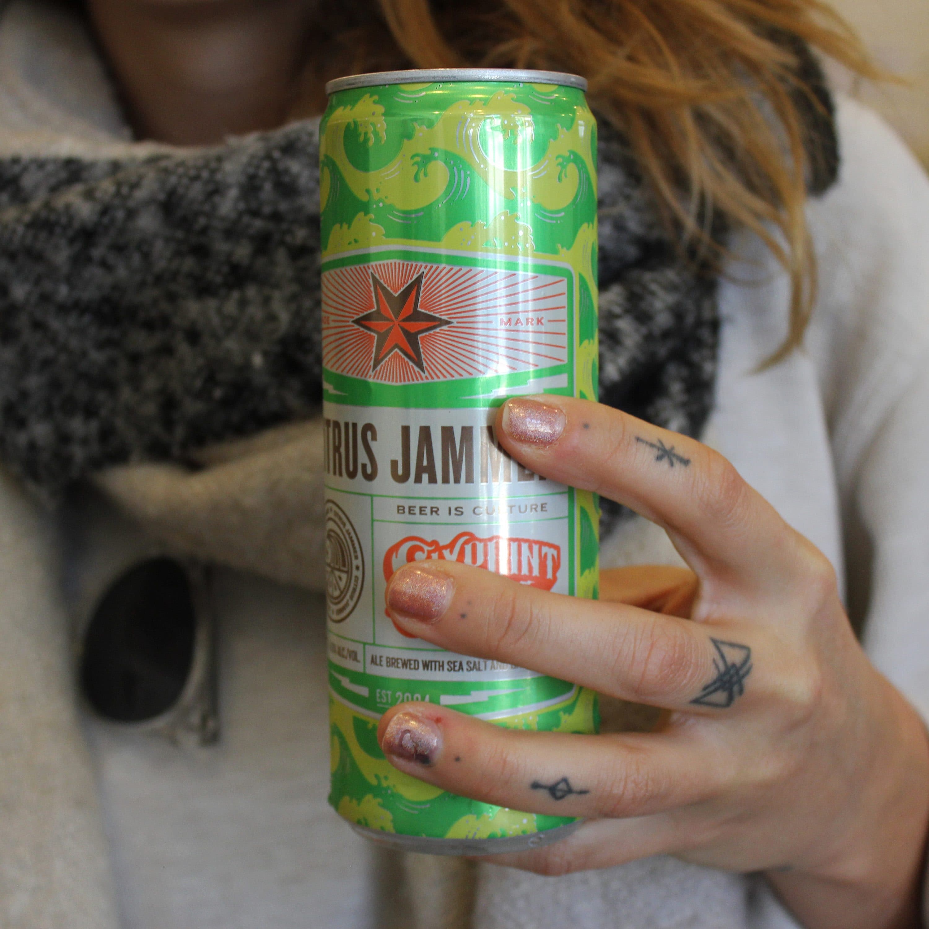 A person with shoulder-length hair holds a can of citrus jam beer covered in a green, yellow, and white design. The person wears a light gray and white sweater with a gray scarf. Their fingers have tattoos and glittery nail polish.