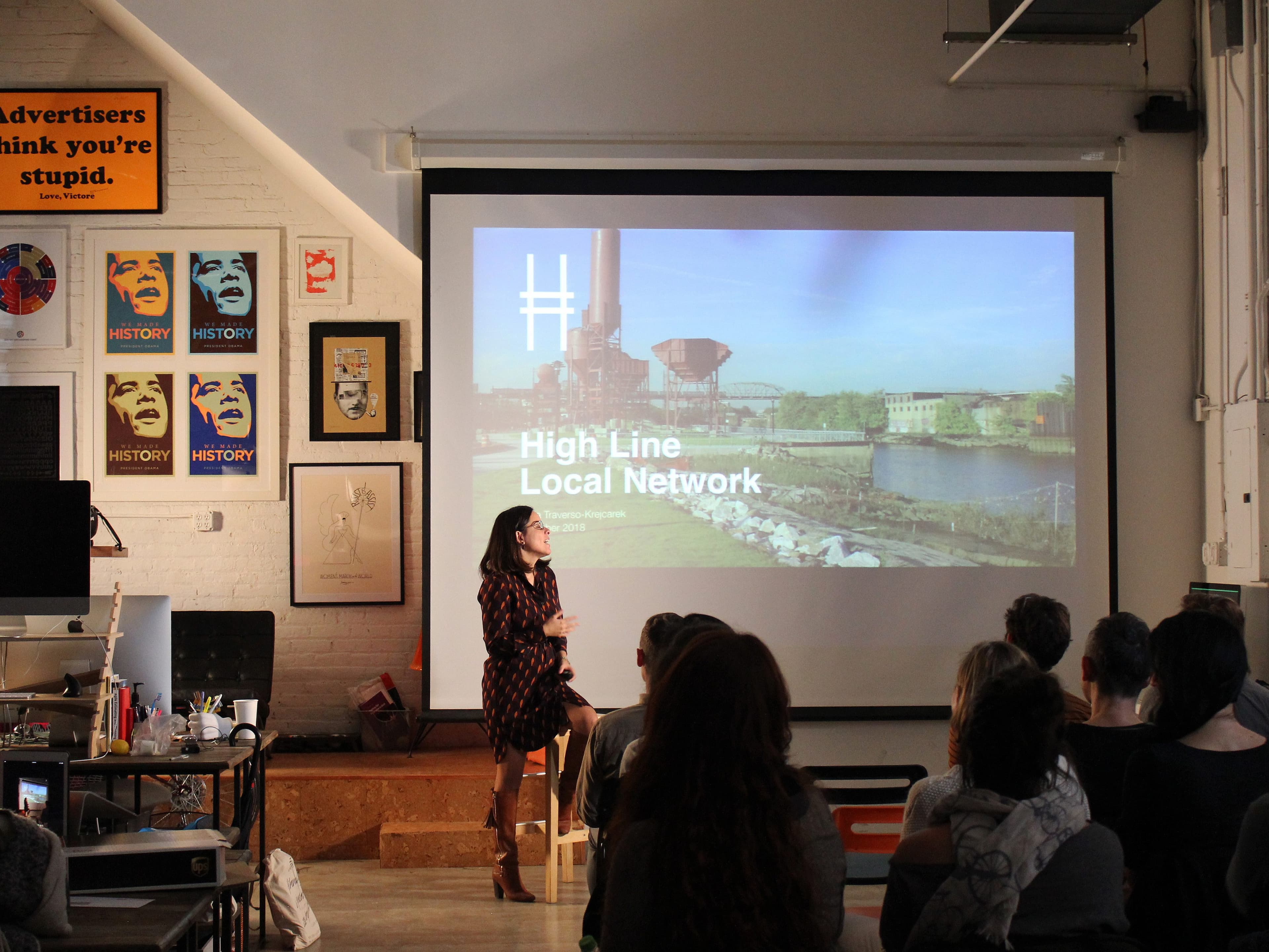 A woman stands in front of a seated audience, presenting a slideshow titled "High Line Local Network". The setting appears to be a creative studio with posters on the wall and bright natural light coming through the windows.