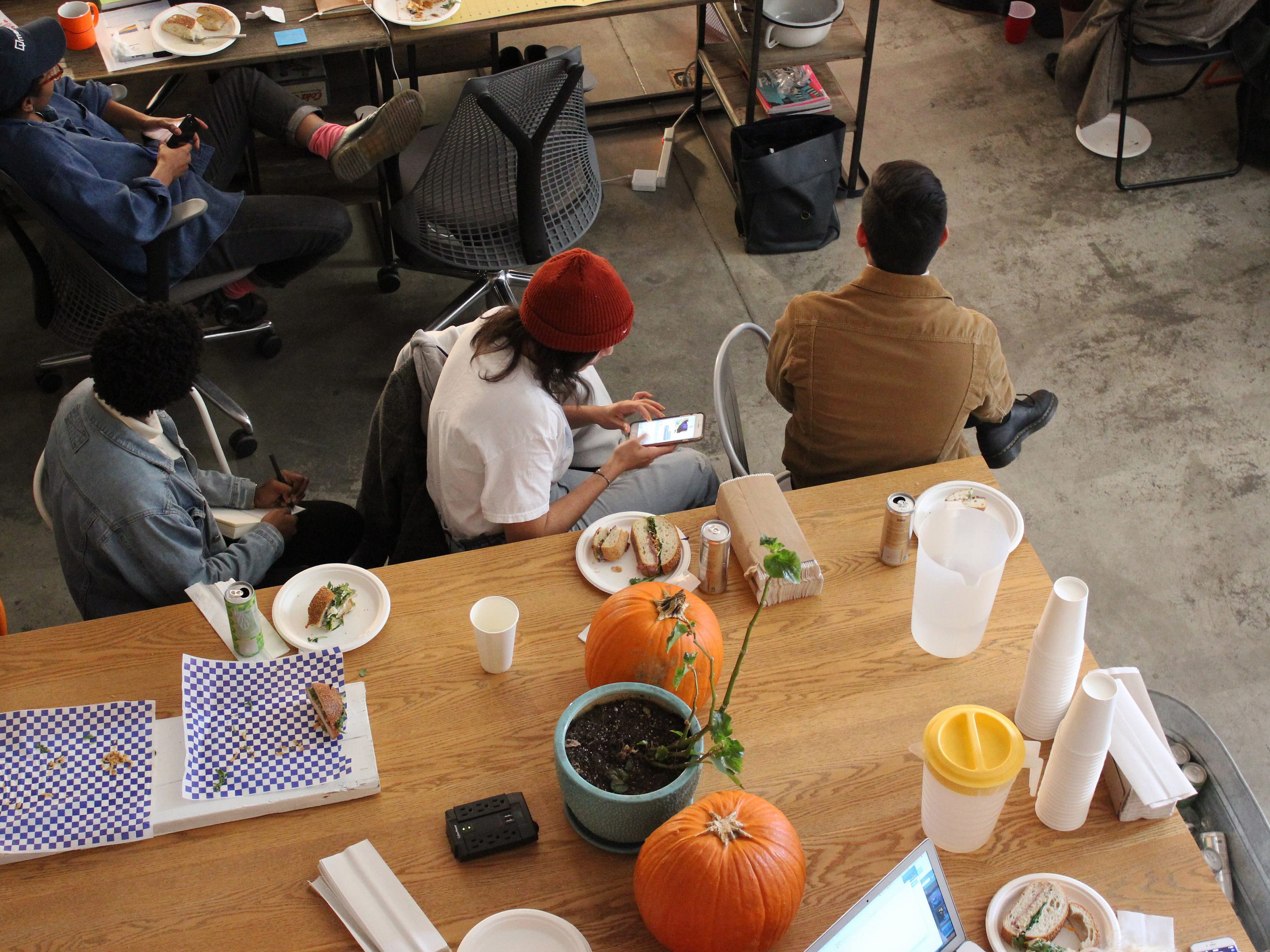 A group of people are seated at a wooden table with pumpkins, food, and drinks, engaging in various activities. Some are using their phones, while others are conversing or eating. The area has a casual, communal atmosphere with plates of food and beverages.