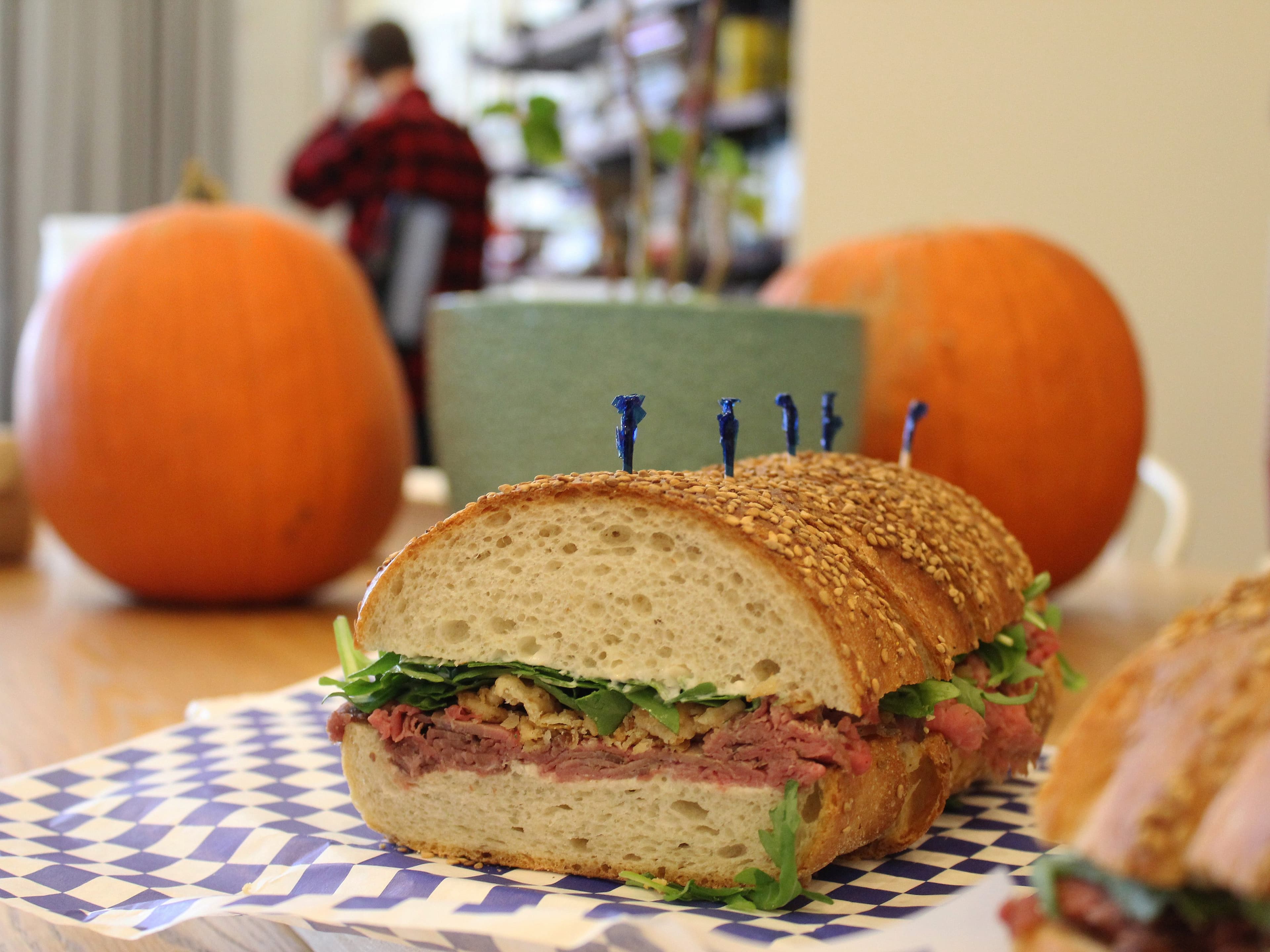 A close-up of a sandwich cut in half, placed on a blue and white checkered paper. The sandwich contains roast beef, arugula, and onions on sesame seed bread. In the background, there are two pumpkins and a blurred person in a red plaid shirt.