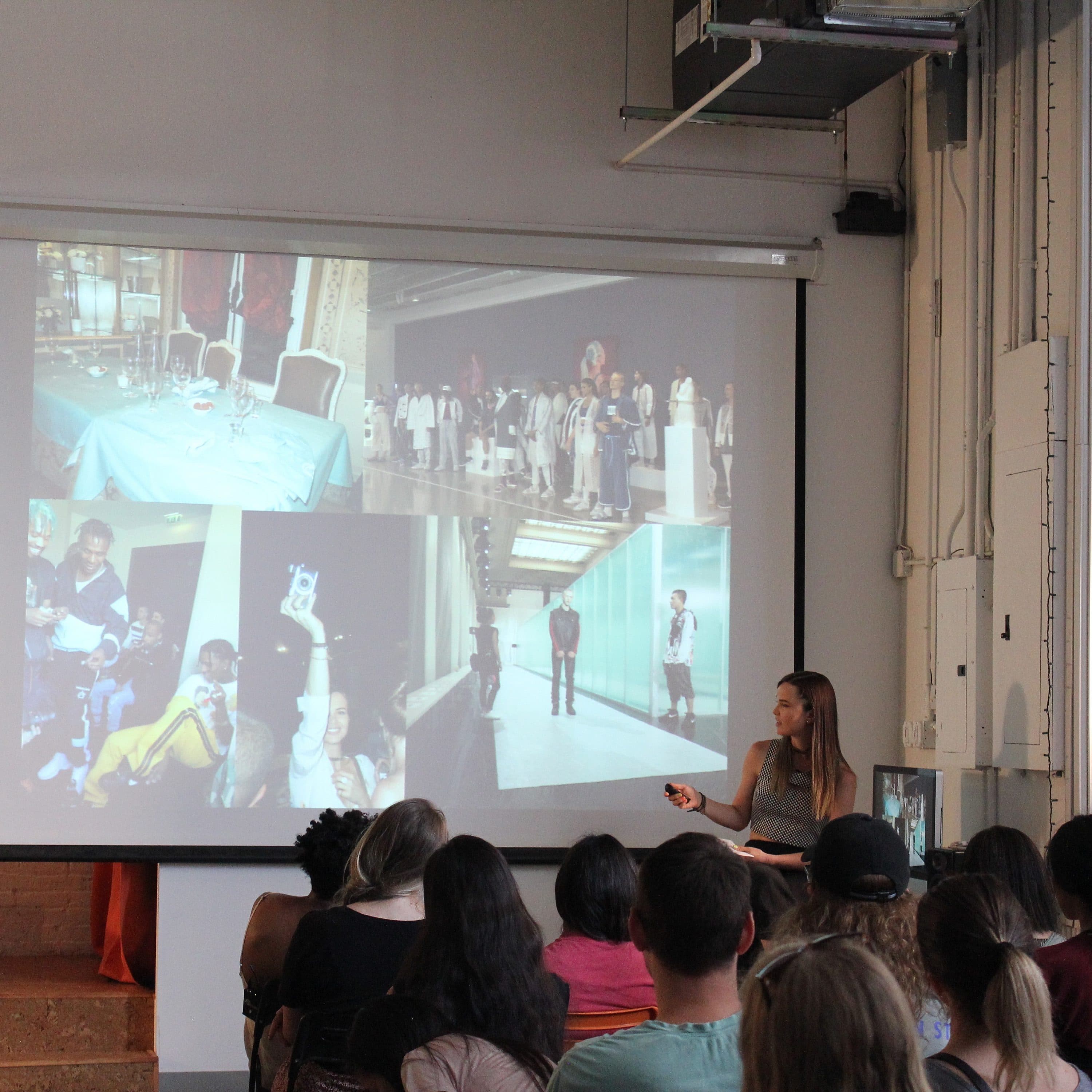 A group of people are seated in an art studio, listening to a woman giving a presentation. She stands next to a large projection screen displaying several fashion and art images. The room has artwork on the walls and various materials and equipment around.