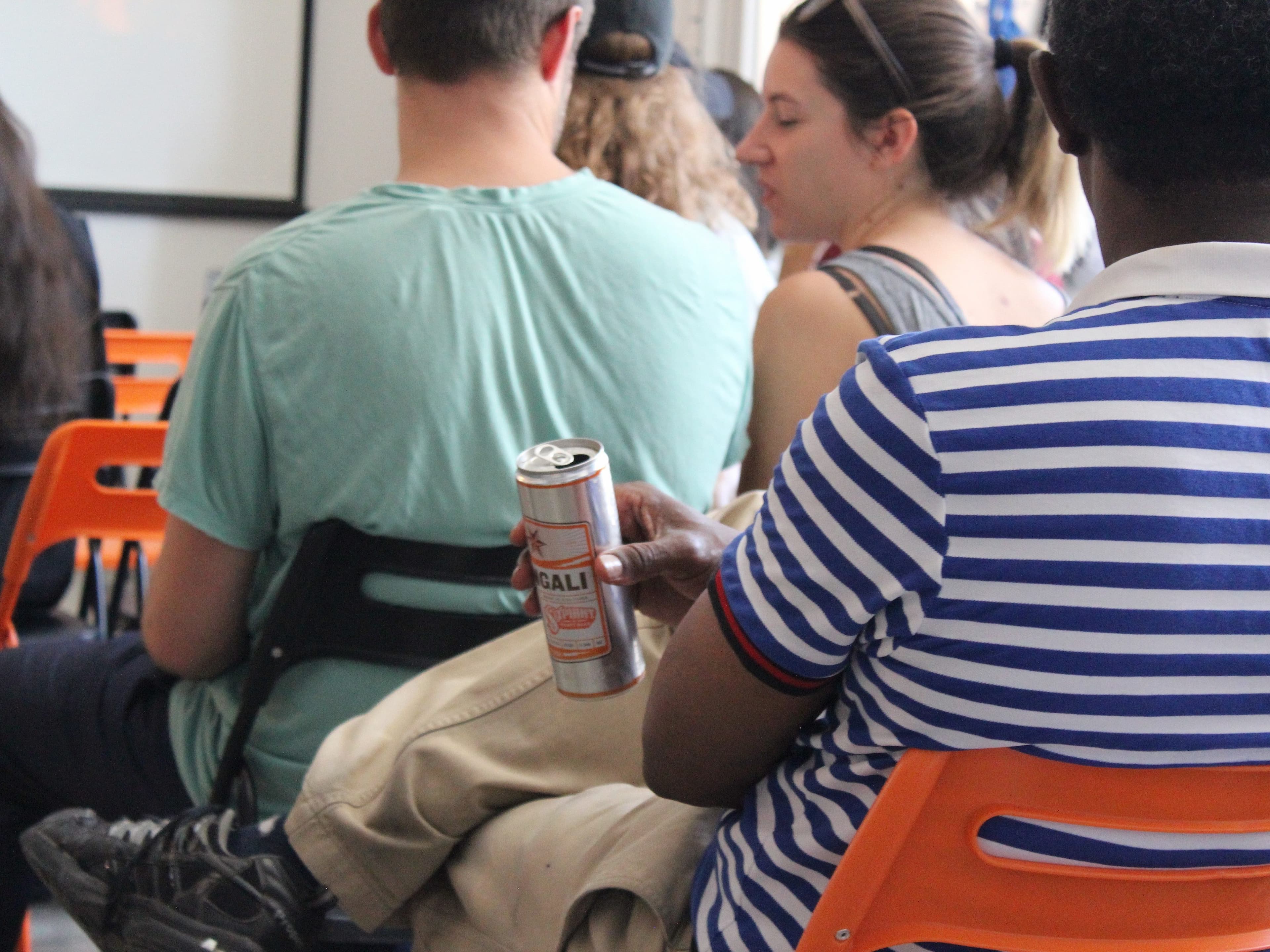 A group of people sitting on orange chairs in a classroom or seminar setting. A person in the foreground wearing a blue and white striped shirt holds an open can of beer. The focus is mainly on the people sitting, with a projector screen visible in the background.
