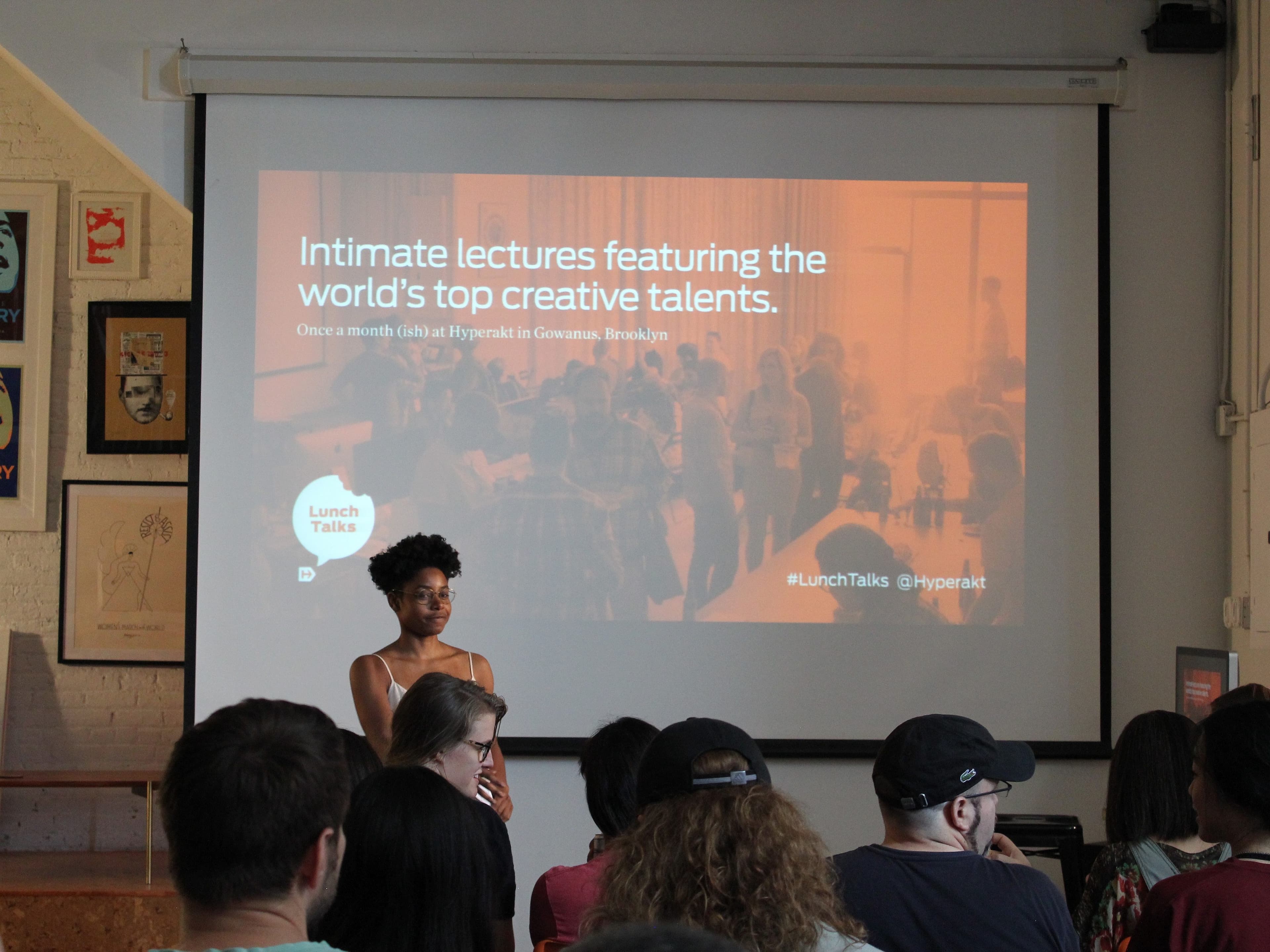 A woman stands in front of a crowd in a room, speaking at an event. Behind her, a screen displays a presentation titled "Intimate lectures featuring the world's top creative talents." The audience is seated and attentively watching the speaker.