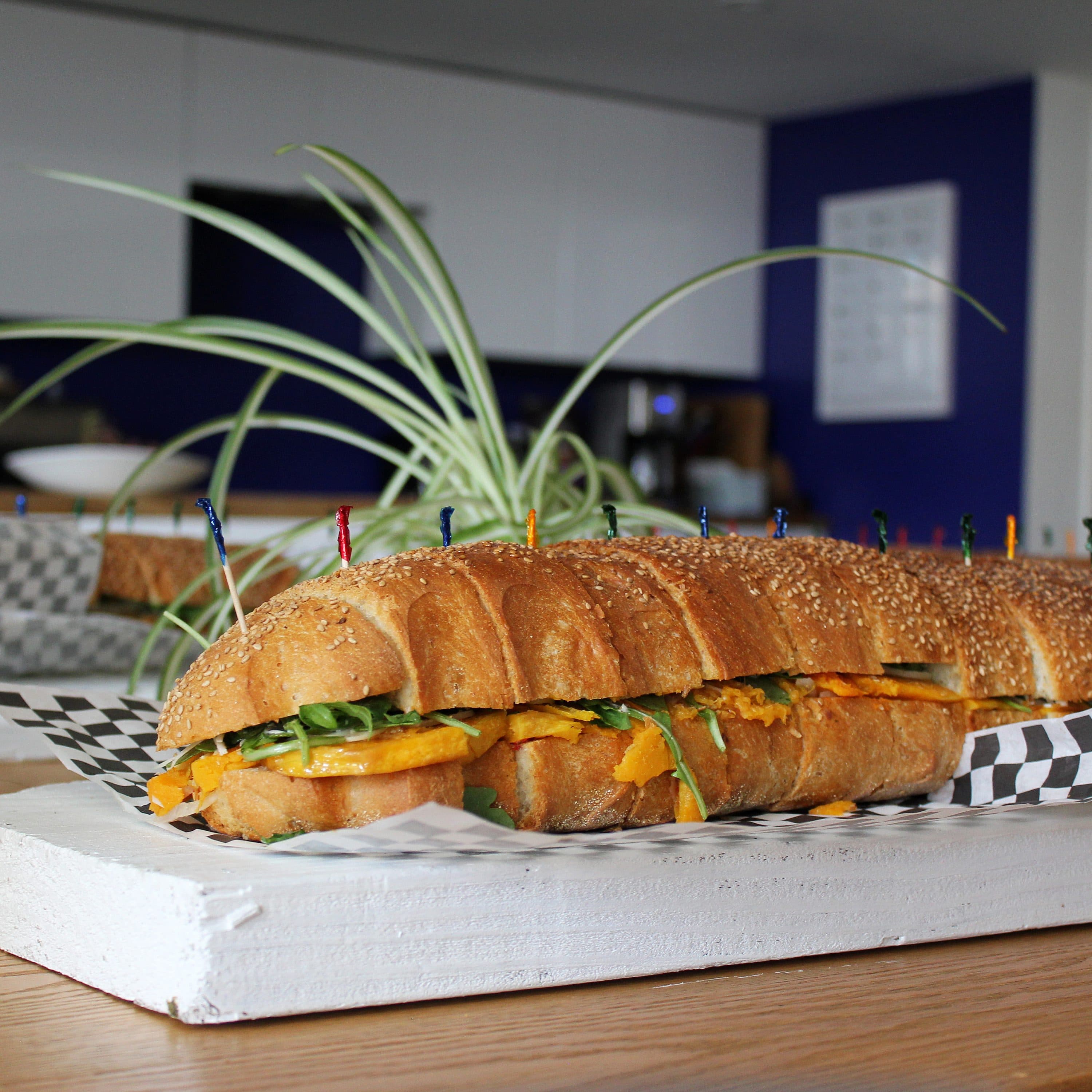 A long sandwich with sesame seed topped bread, filled with various ingredients including greens and cheese, is placed on a wooden tray lined with checkered paper. The background features a modern kitchen with a plant in focus.