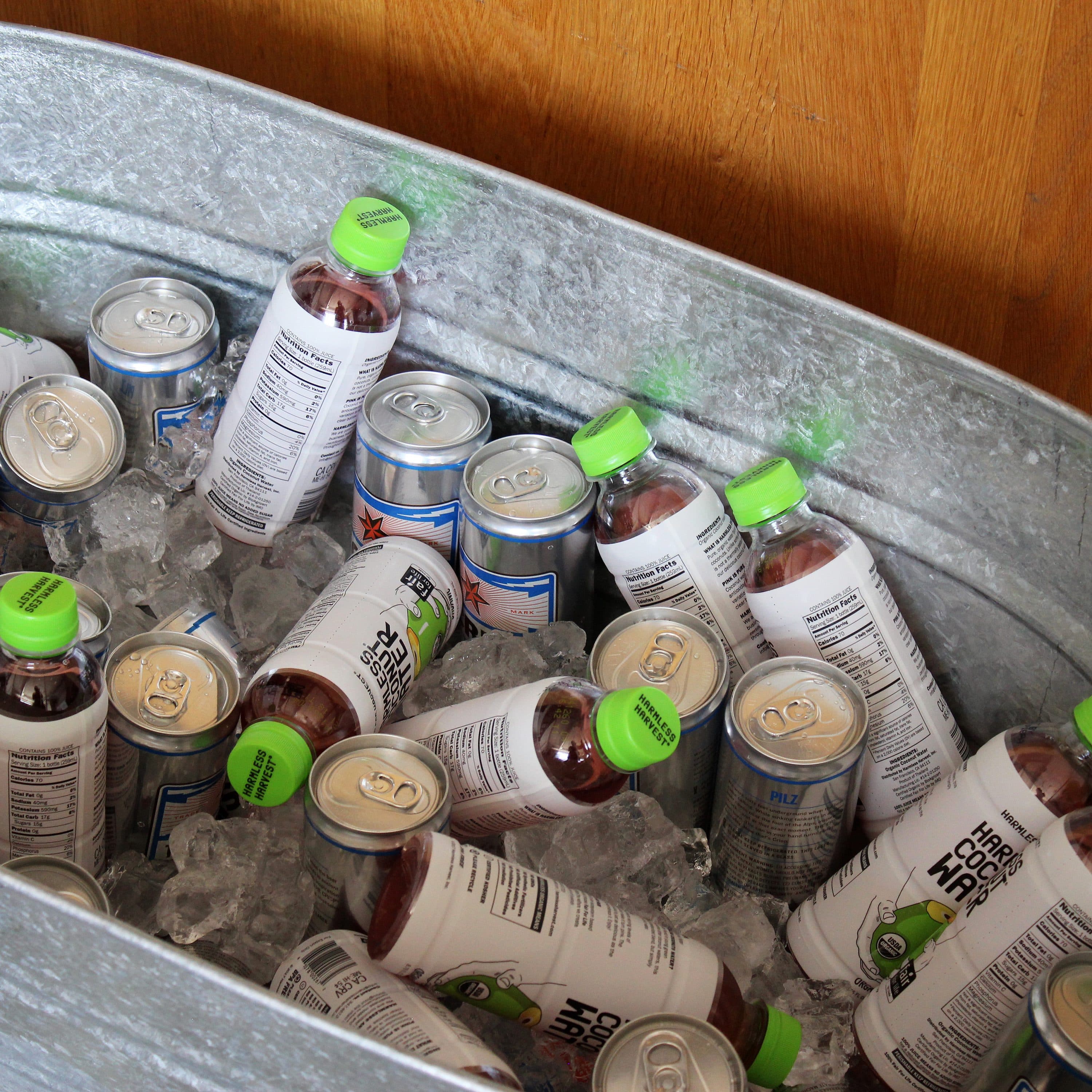 A large metal tub filled with ice, plastic bottles with green caps, and cans of sparkling water. The bottles and cans are chilled, ready for serving. The background features a wooden surface.