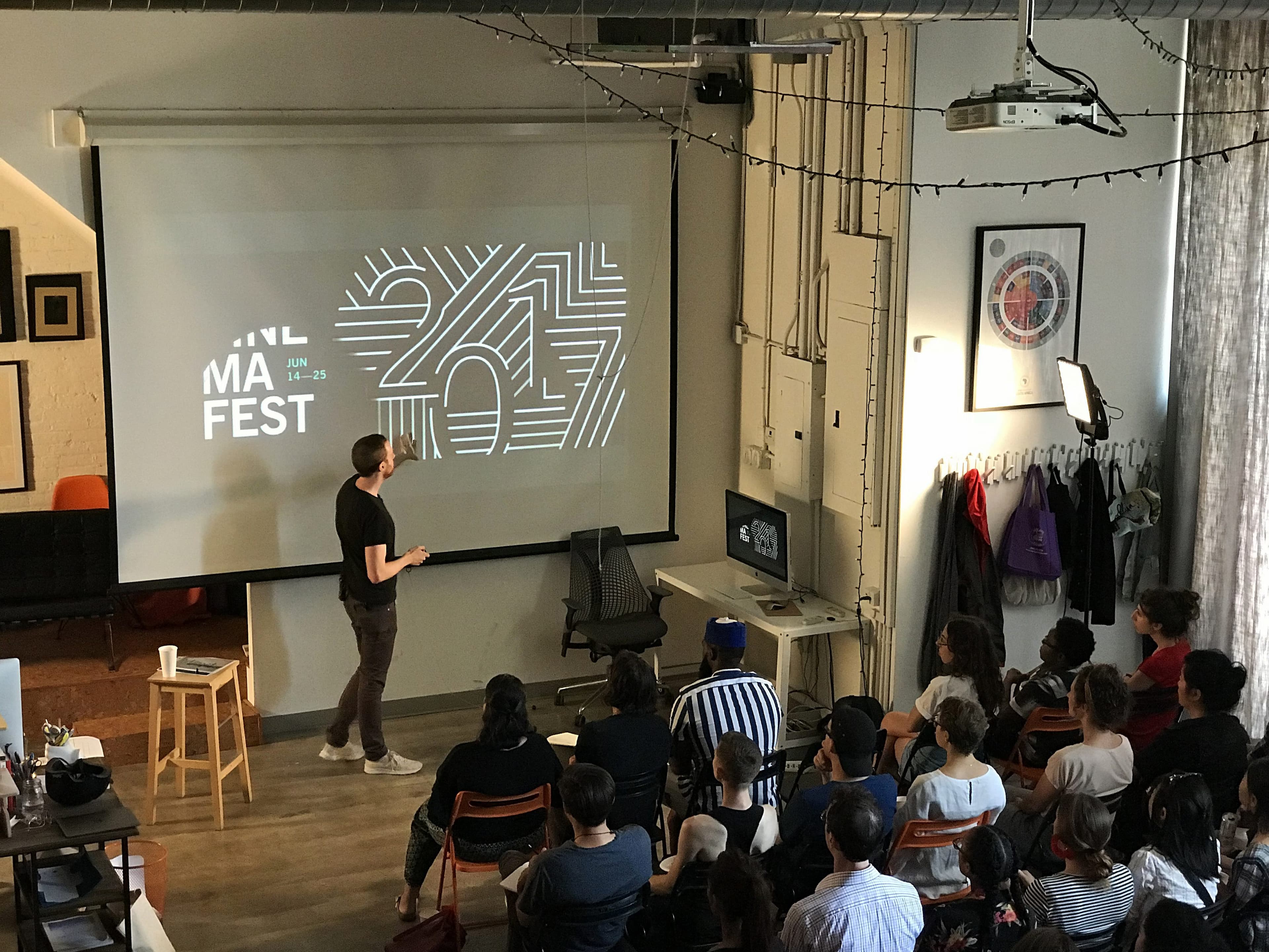A person stands in front of an audience presenting slides projected on a screen that reads “ANIMA FEST 2017”. The room is dimly lit, with string lights on the ceiling and various framed pictures on the walls. The audience is seated and attentive.