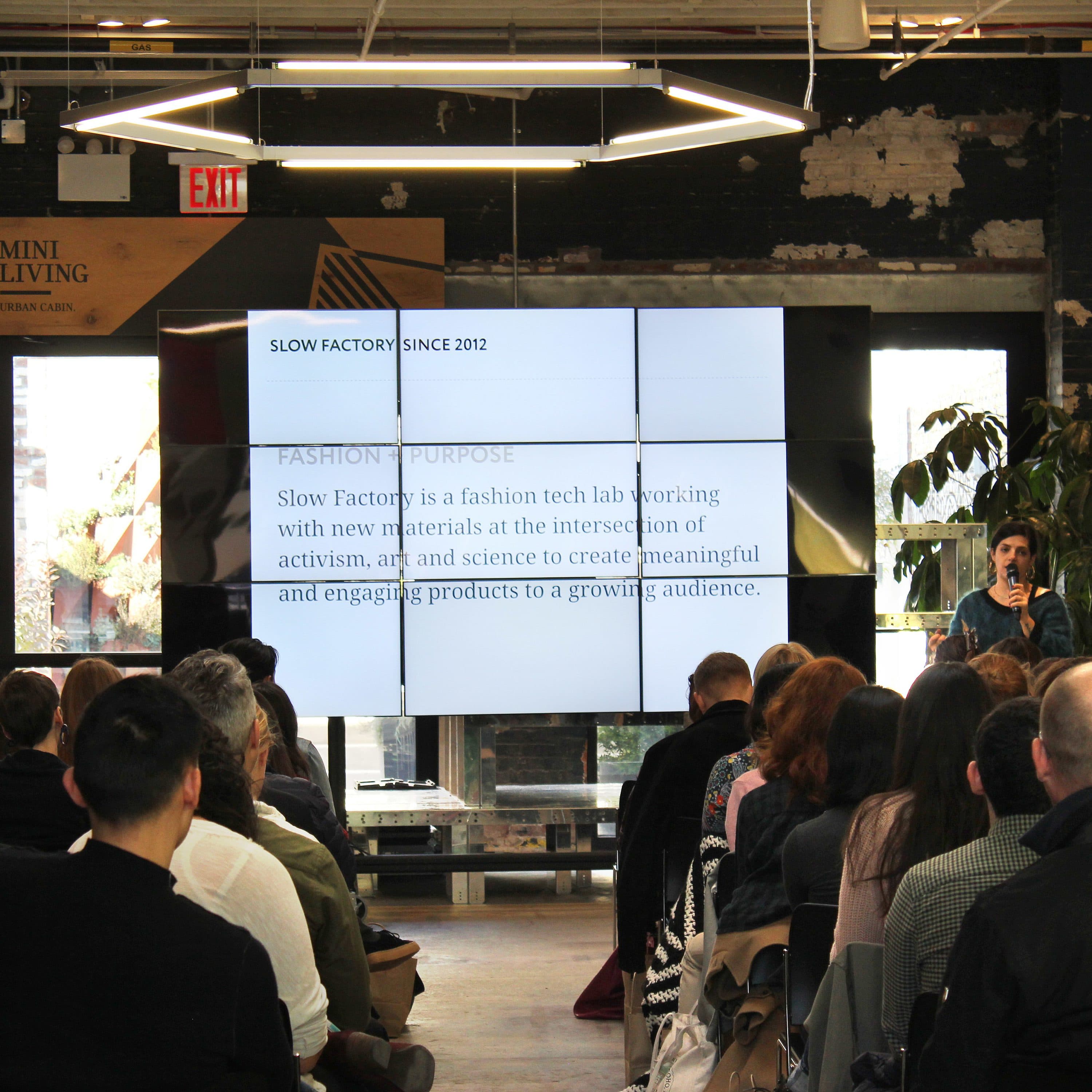 A speaker stands near a large screen with a presentation titled "SLOW FACTORY SINCE 2012," addressing an audience in an industrial-style room. The audience, seated in rows, listens attentively. The room features exposed brick walls and lighting fixtures.