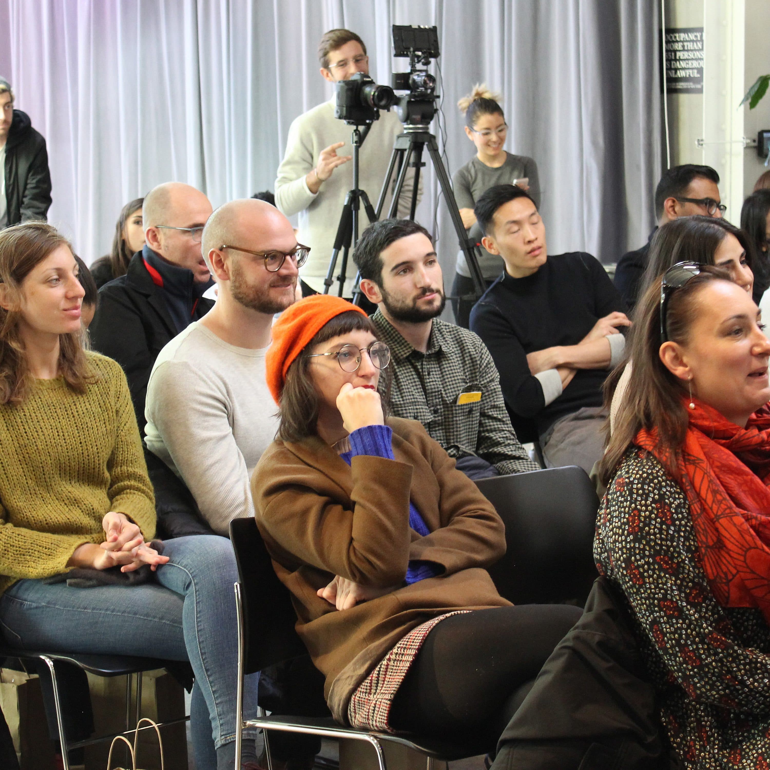A diverse group of people are seated and attentively watching something out of frame in a well-lit room. Some are smiling or deep in thought. There is a person with a camera on a tripod in the background and a large plant to the right. The atmosphere appears casual and engaged.