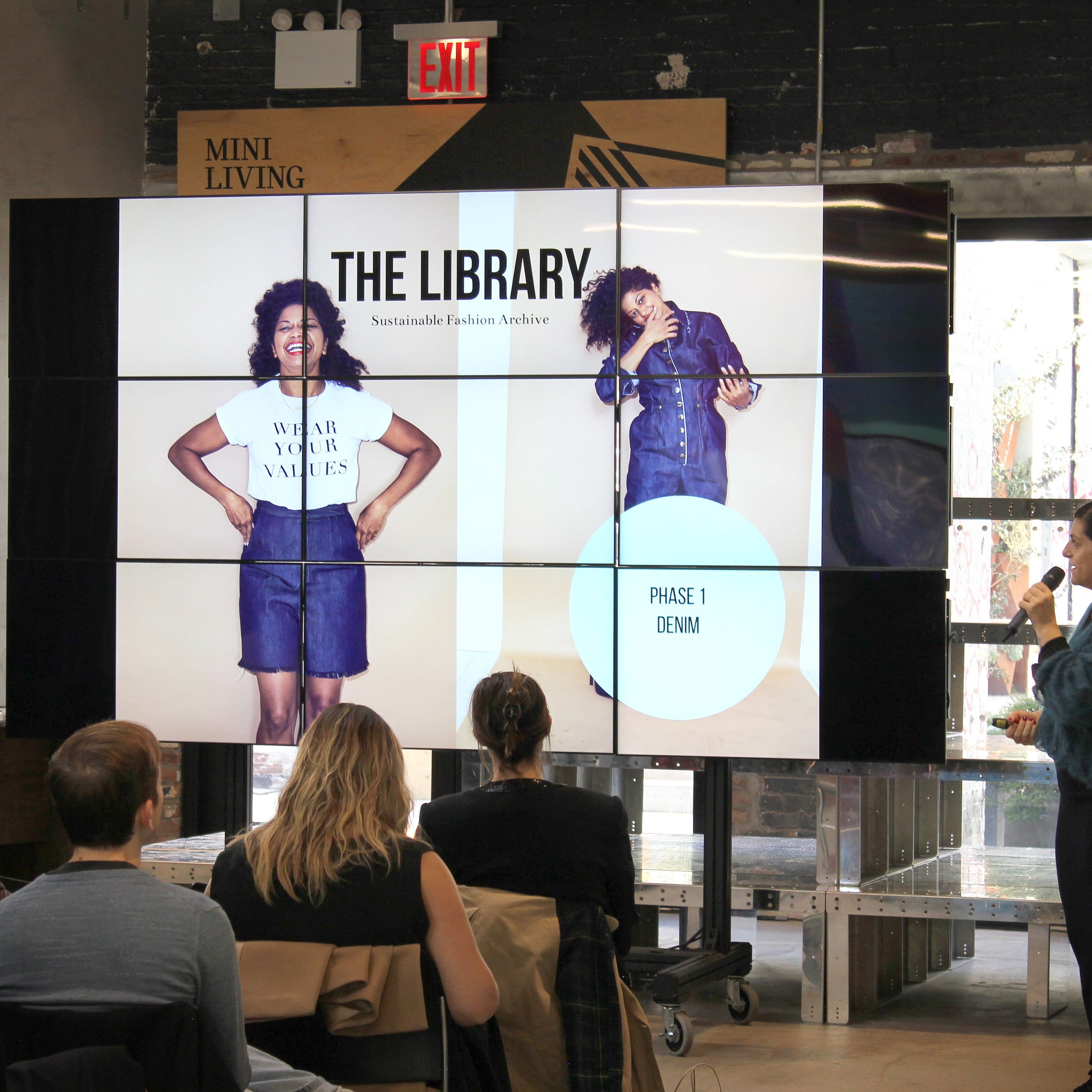 A woman is presenting in front of an audience with a microphone, pointing to a large screen displaying an image of a person wearing a denim outfit. The screen reads: "The Library: Sustainable Fashion Archive" with the text "Phase 1 Denim." The audience is seated and attentive.