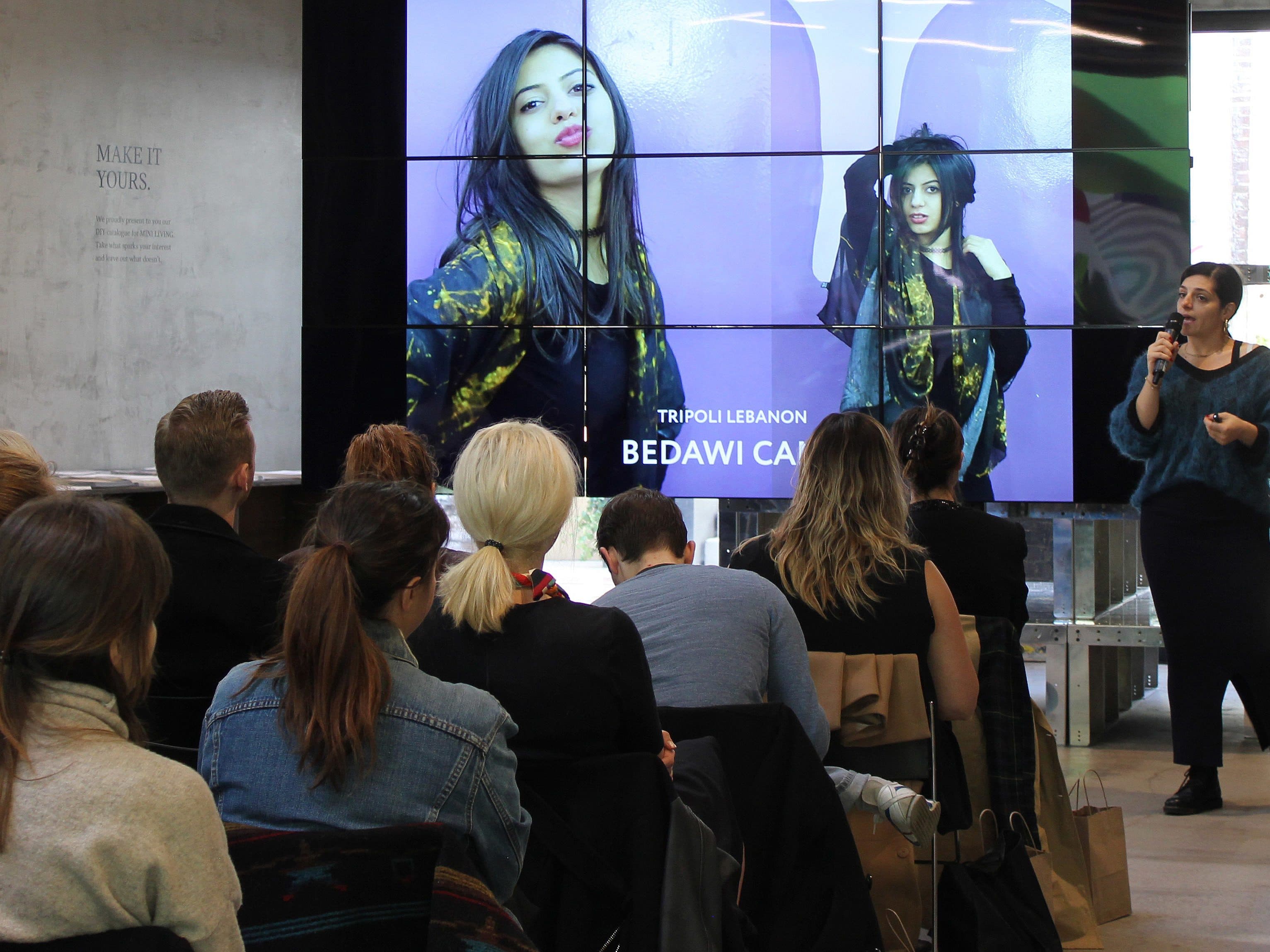 A woman is speaking into a microphone in front of an audience seated in a modern indoor space. Behind her is a large screen displaying images of another woman posing, labeled "Tripoli Lebanon Bedawi Camp." The audience appears to be focused on the presentation.