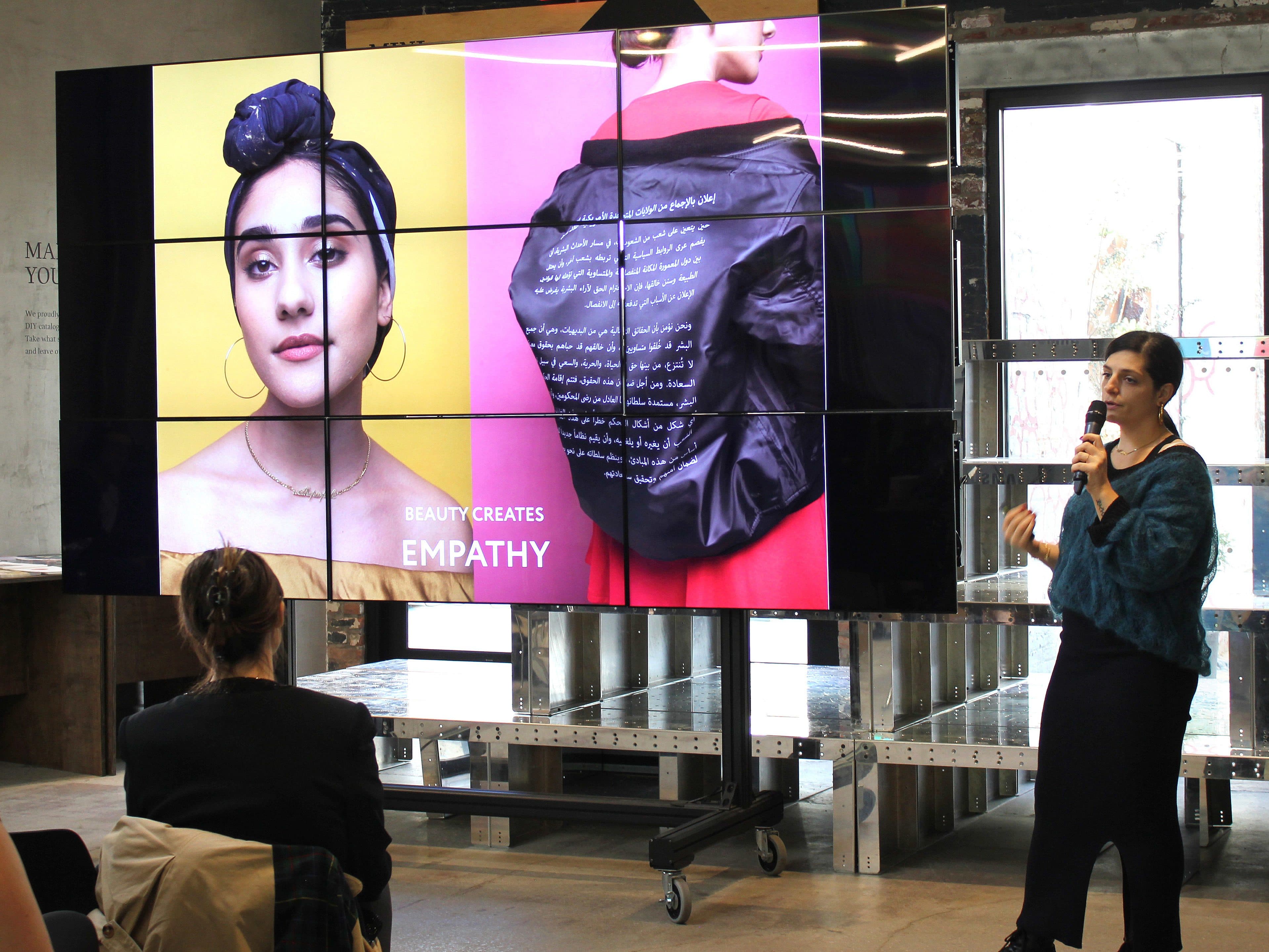 A woman presents on stage with a microphone, standing next to a large screen displaying two fashion photos and the text "BEAUTY CREATES EMPATHY." The audience is seated, and the setting appears to be an art gallery or modern presentation space.
