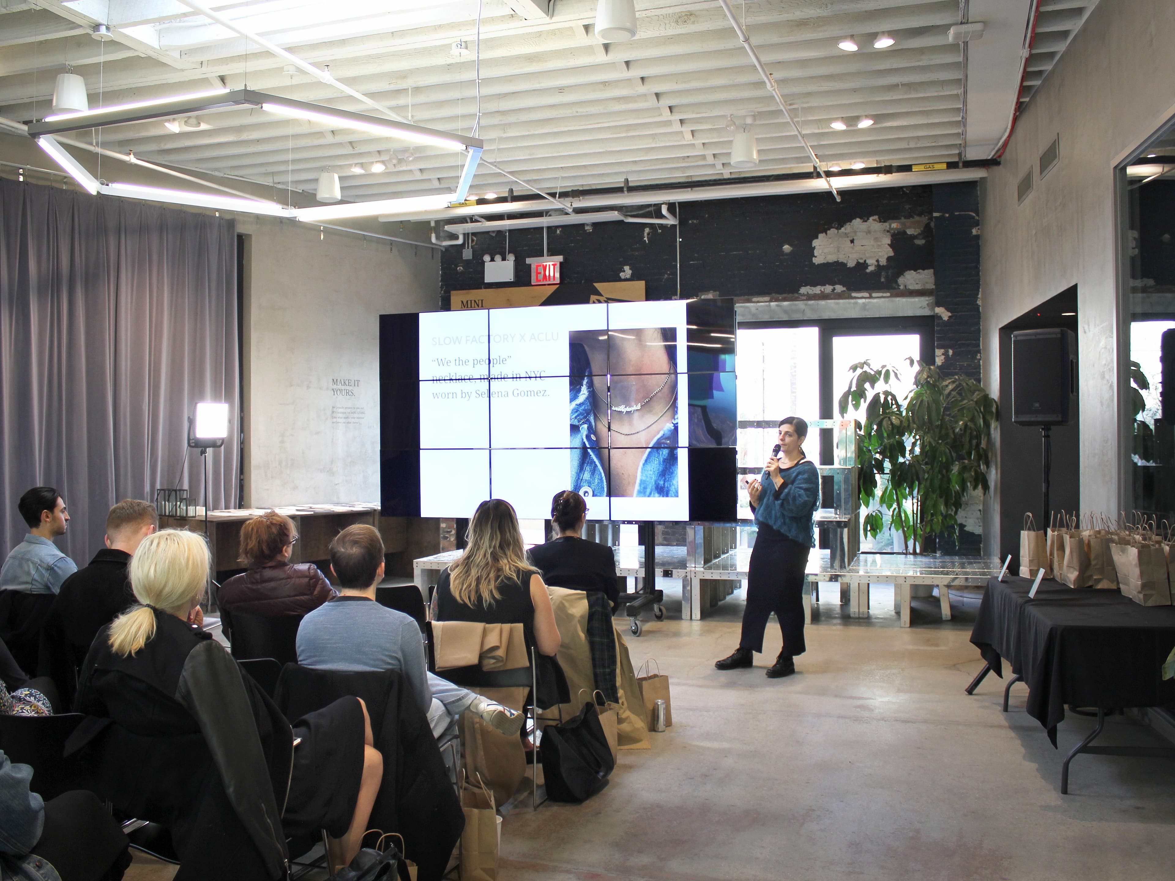 A speaker presents to an audience in a modern, industrial-style room. The audience is seated and facing a large screen displaying a jewelry ad. Tables with gift bags are placed on the right side of the room. The room has large windows, exposed brick, and high ceilings.