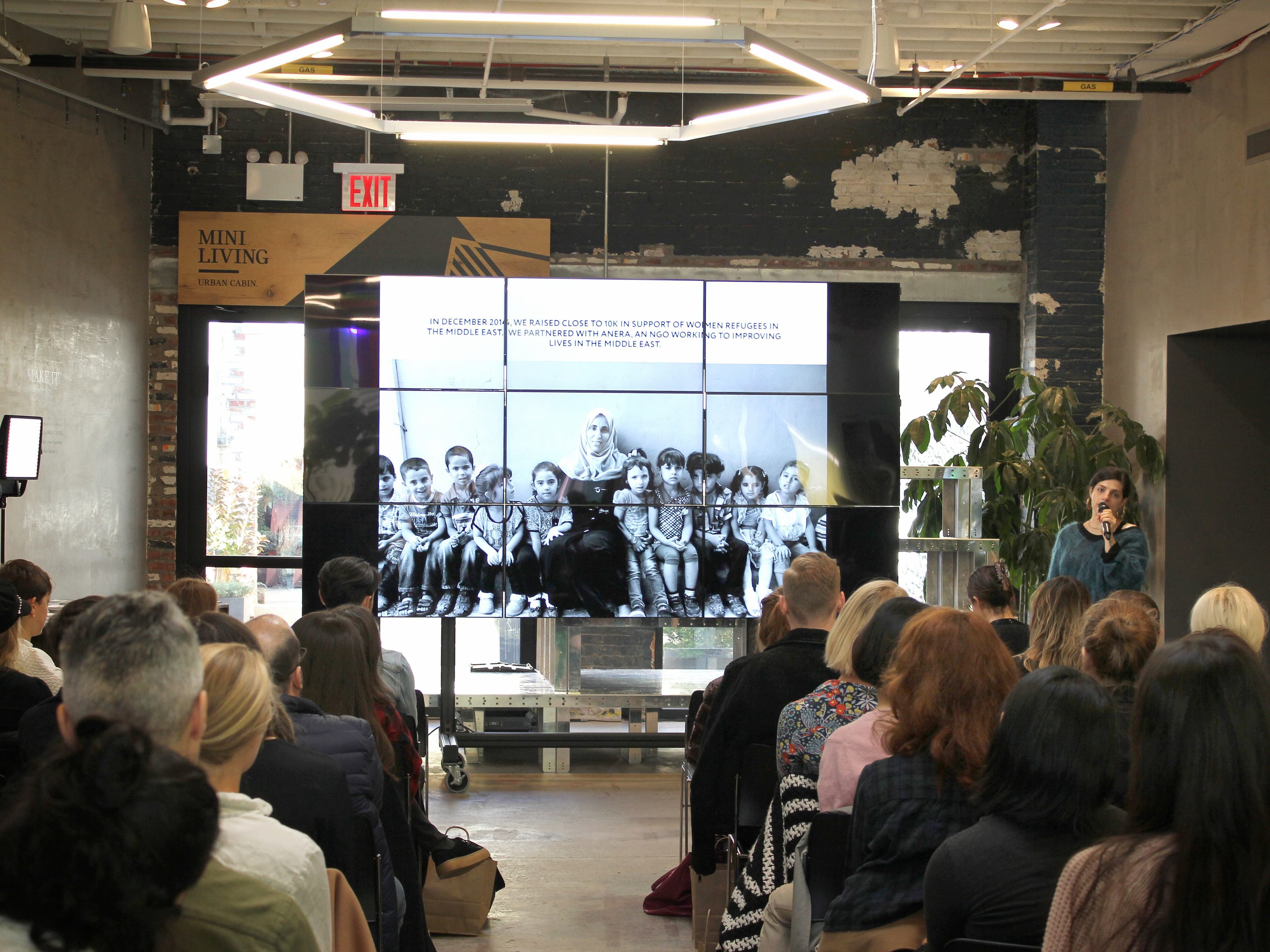 A group of people sit in chairs facing a large screen, listening to a speaker. The screen shows a black and white photo of children sitting together. The event takes place in an industrial-style room with brick walls and overhead lighting.