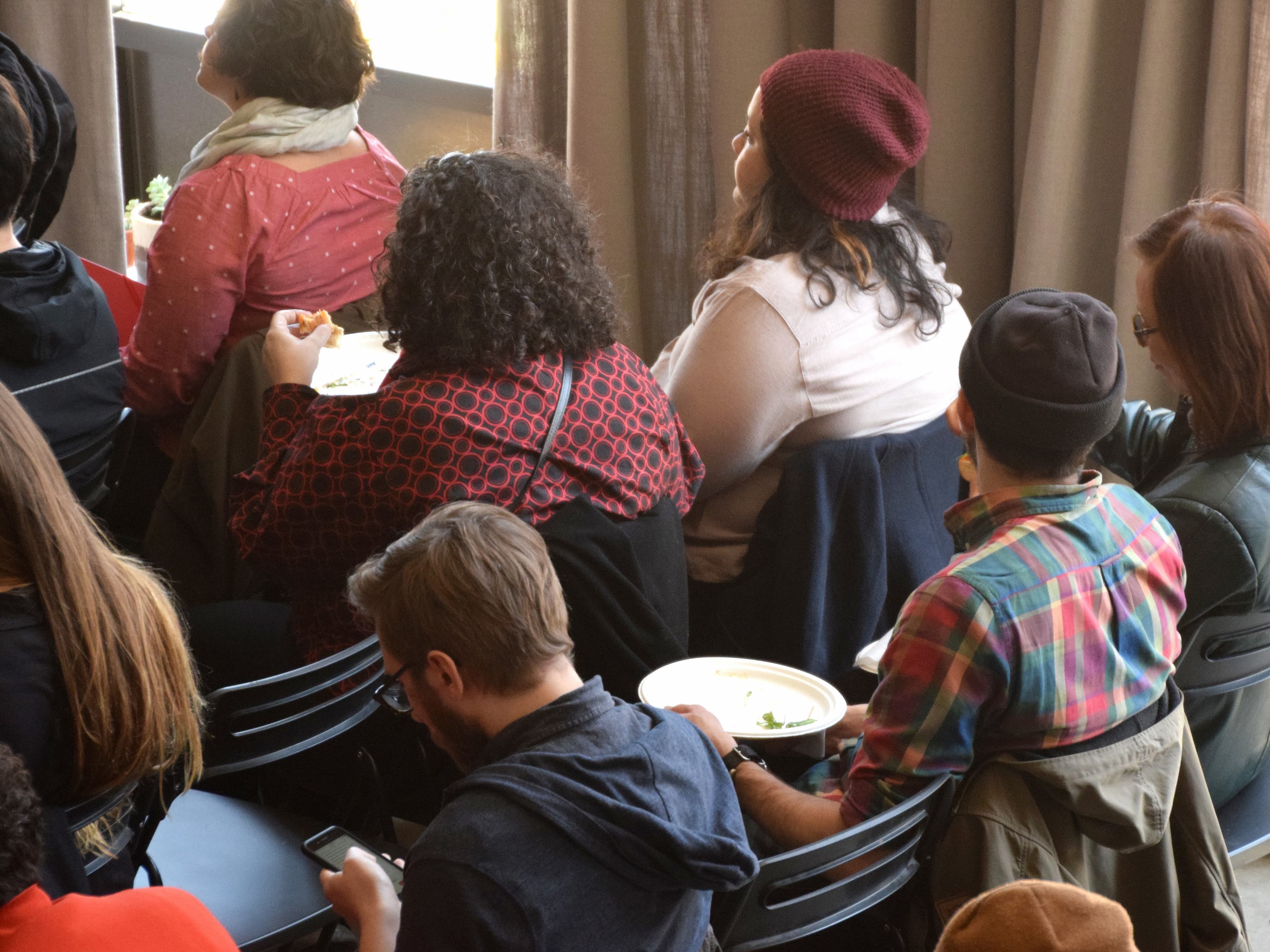 A group of people are seated closely together in chairs, facing away from the camera. Some are eating off plates, others are looking at their phones. One person wears a red beanie, another a black beanie, and multiple others have jackets on the backs of their chairs.
