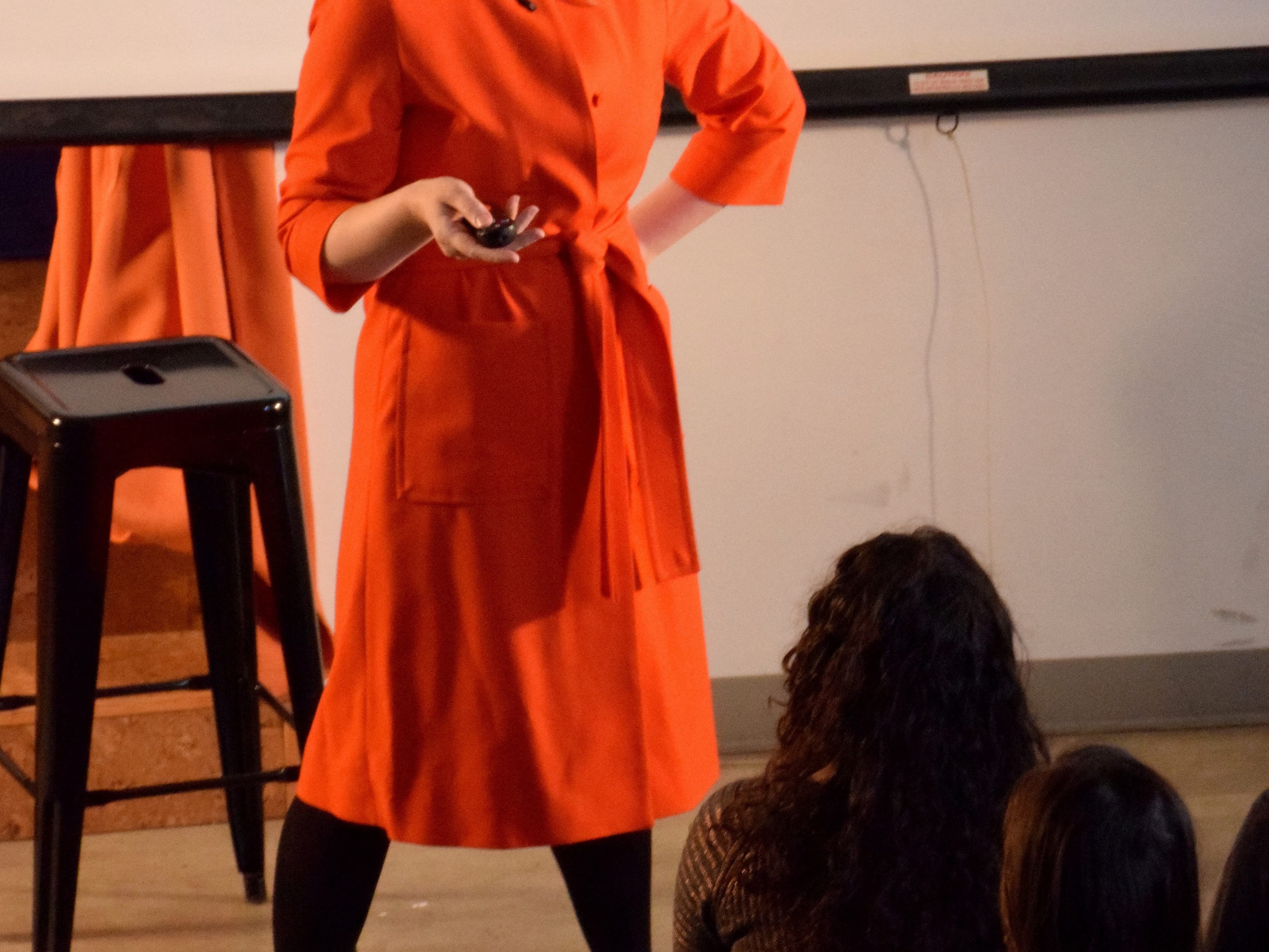 A woman in an orange dress is giving a presentation. She appears to be speaking passionately, holding a small device in one hand, with the other hand on her hip. She is standing in front of an audience seated indoors, with a stool and a projected screen behind her.