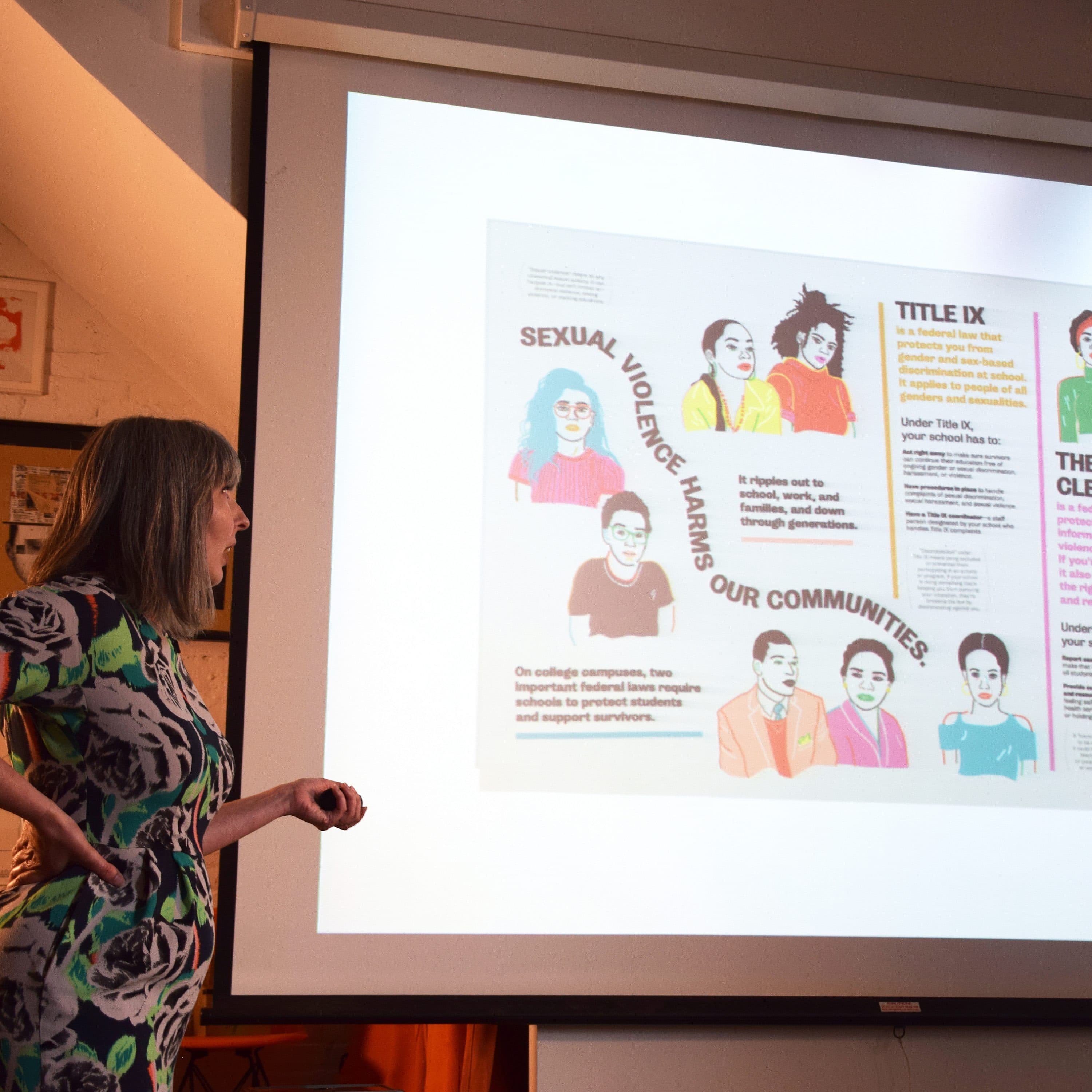 A person with long gray hair and a patterned dress is standing in front of a screen displaying educational posters about sexual violence, community support, and Title IX. The screen features cartoon-style illustrations and informative text on these topics.
