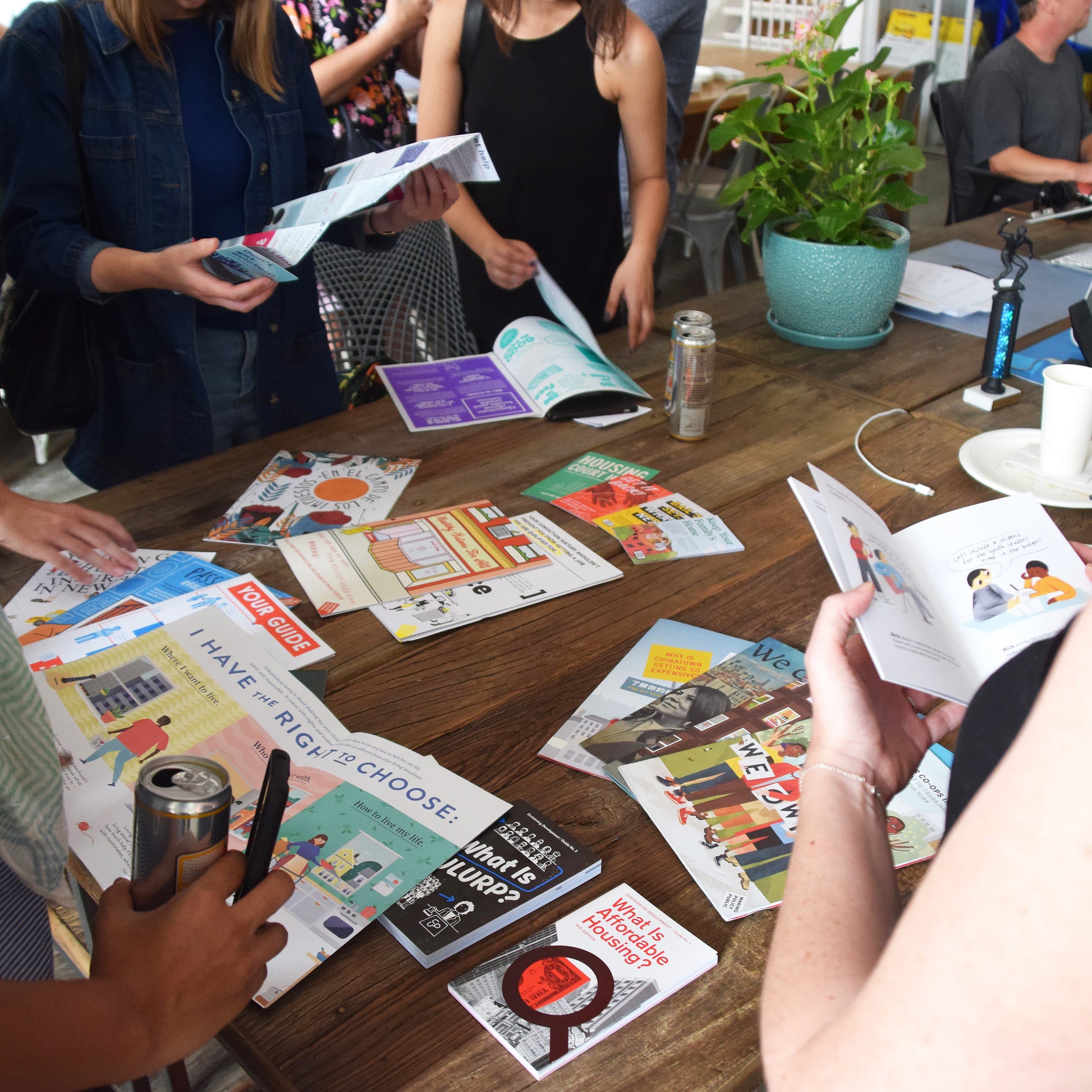 A group of people stand around a table covered with various pamphlets, leaflets, and magazines. Some individuals are reading through the materials, while others are looking or reaching for items. A potted plant and a candle are on the table among the clutter.