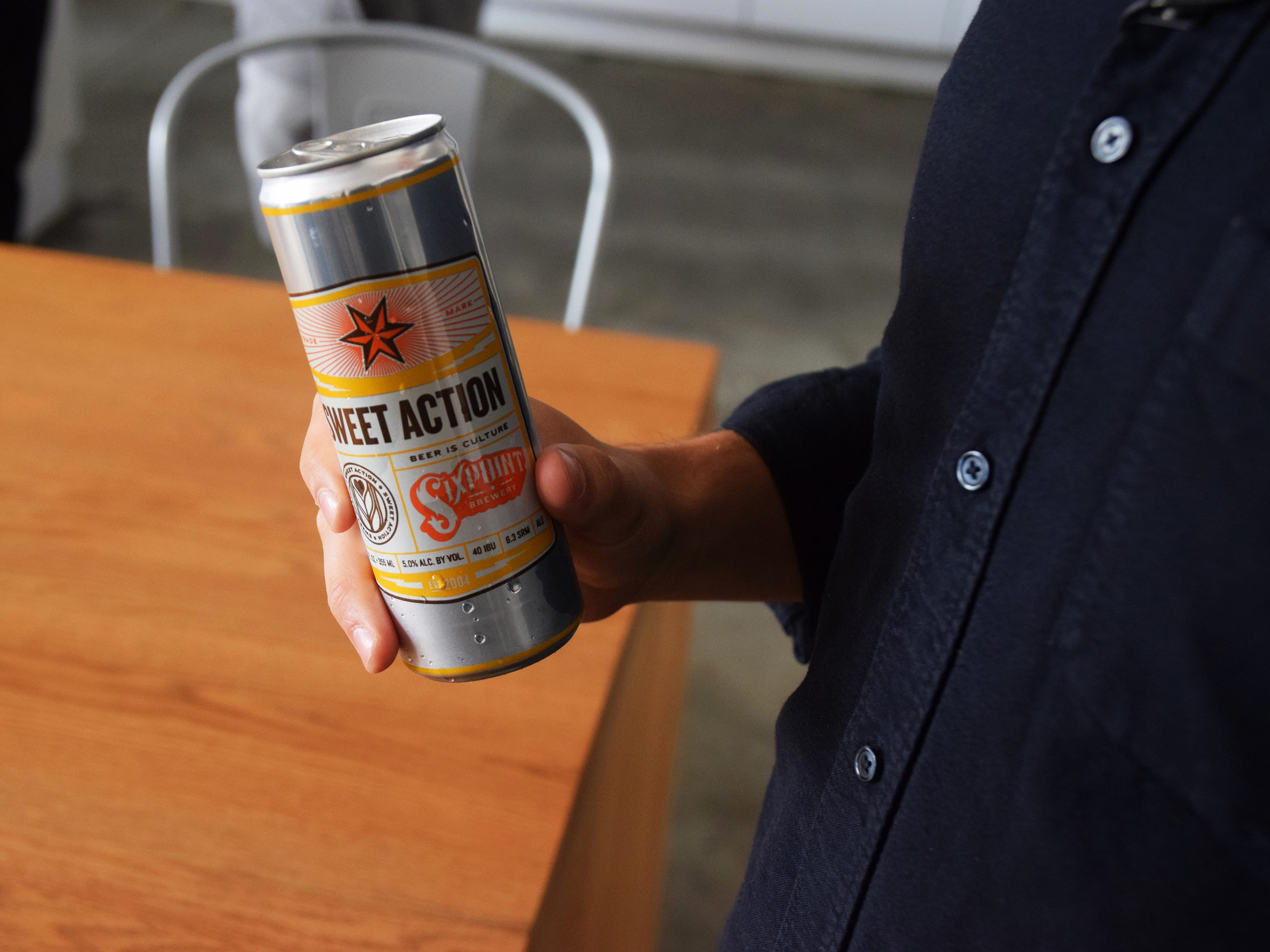 A person is holding a can of Sixpoint Brewery's Sweet Action beer. The can features a star logo and yellow, white, and orange design elements. The person is wearing a dark-colored shirt, and part of a wooden table is visible in the background.