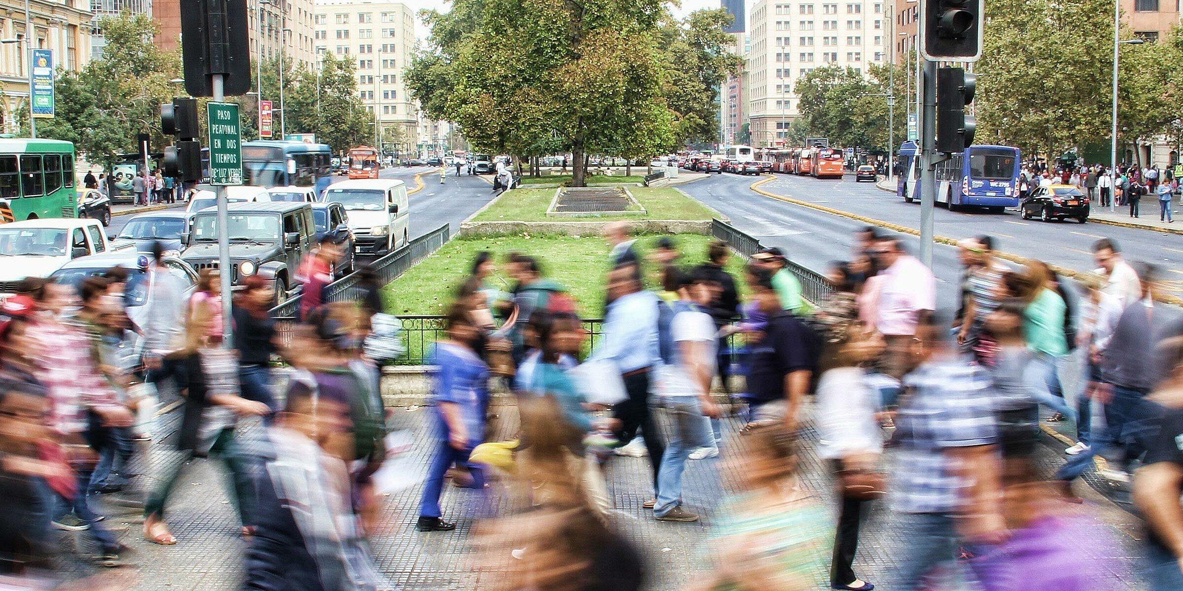 A busy urban scene showing numerous people walking across a crosswalk, with buildings, cars, and trees in the background. The blurred motion of the pedestrians indicates their hurried pace. Traffic lights and a tall tower are visible in the distance.