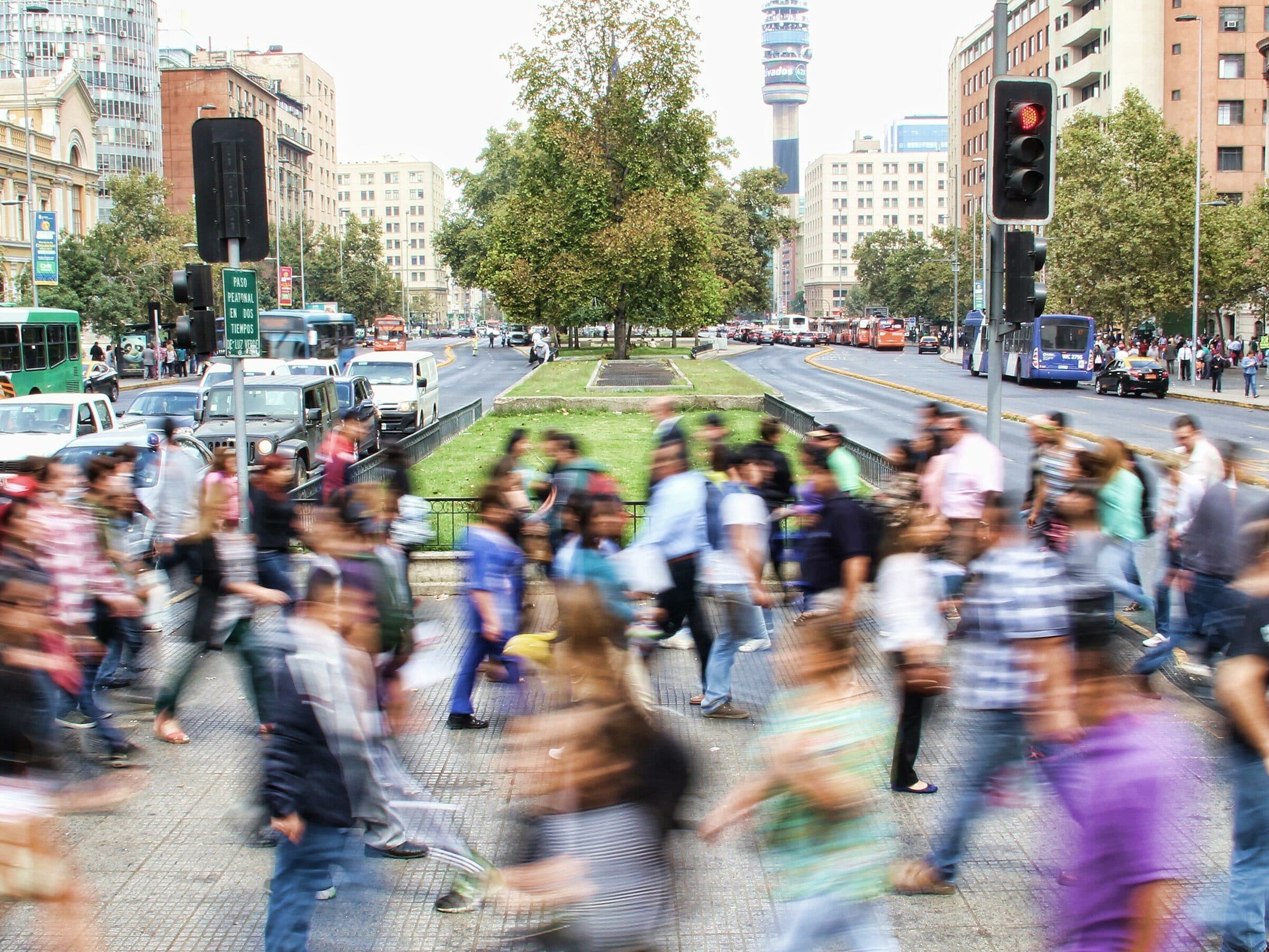 A busy urban scene showing numerous people walking across a crosswalk, with buildings, cars, and trees in the background. The blurred motion of the pedestrians indicates their hurried pace. Traffic lights and a tall tower are visible in the distance.