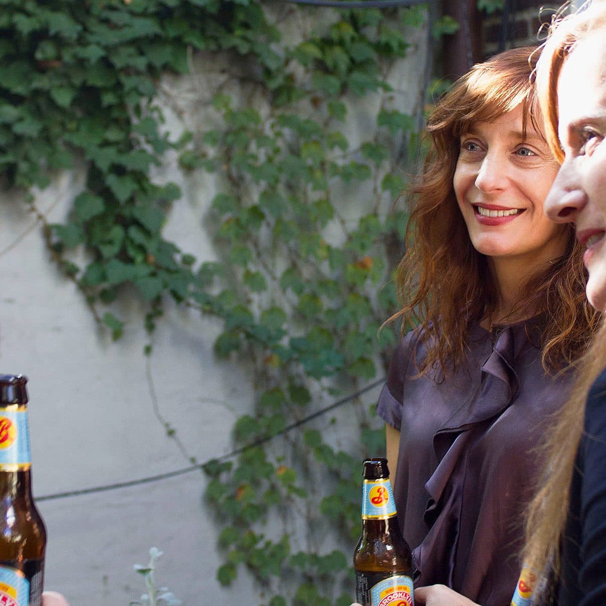 Two women are smiling and holding beer bottles, engaging in conversation outdoors. One woman has long wavy hair and wears a dark top, while the other has long straight hair. The background features a wall covered in ivy and greenery.