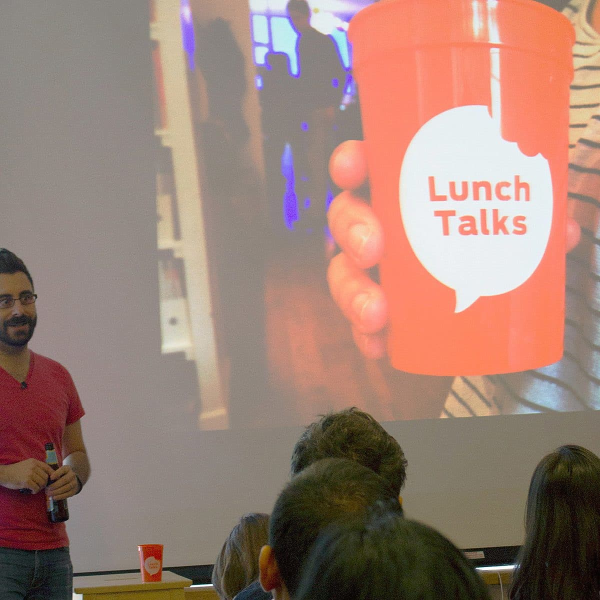A speaker in a red shirt is holding a drink and presenting in front of an audience. Behind the speaker, a large projection displays a close-up of a red cup with the words "Lunch Talks" on it. Several people in the audience are watching attentively.