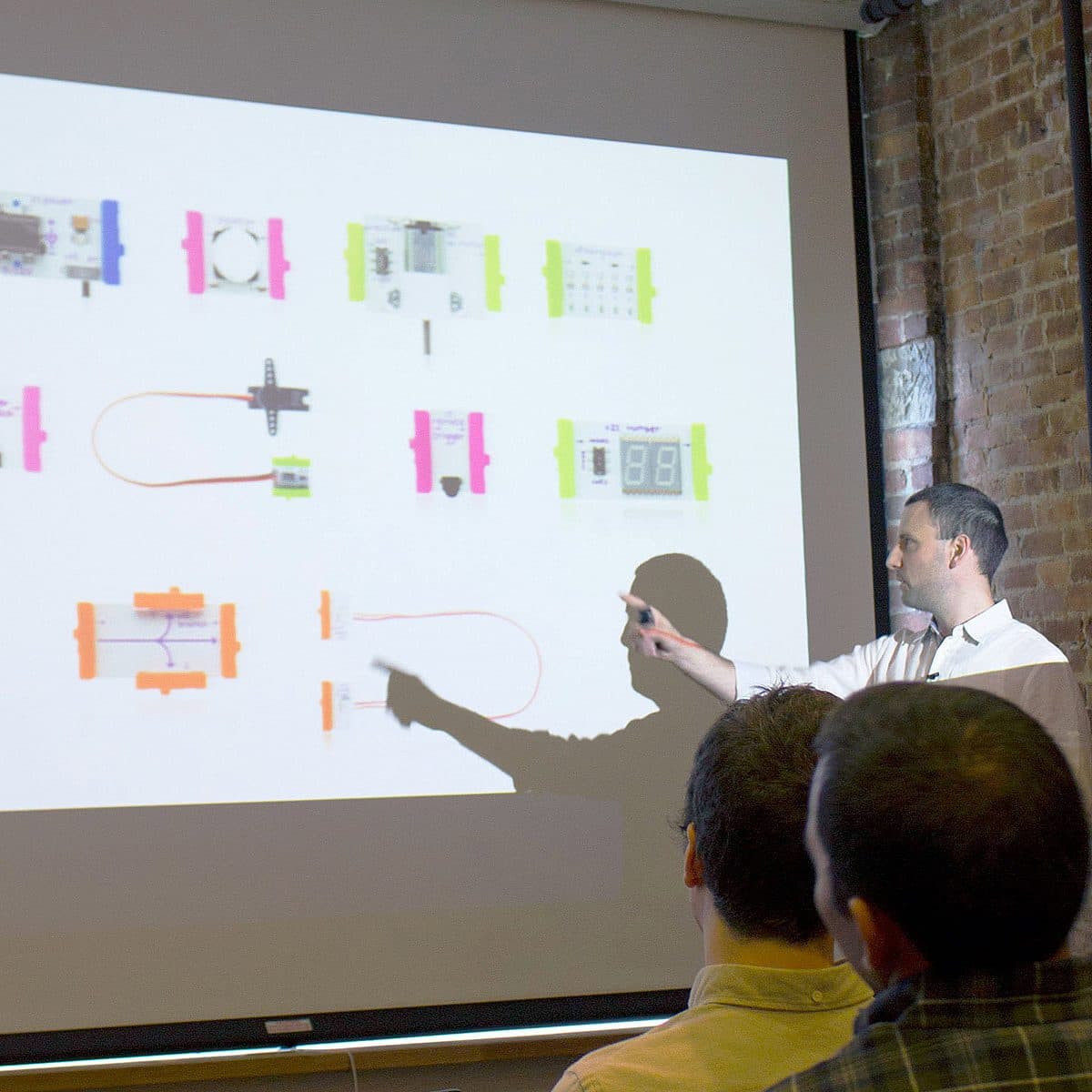 A presenter is pointing at a slide displaying diagrams of electronic components labeled "VCR" in front of a brick wall. The audience is seated and attentively watching the presentation.