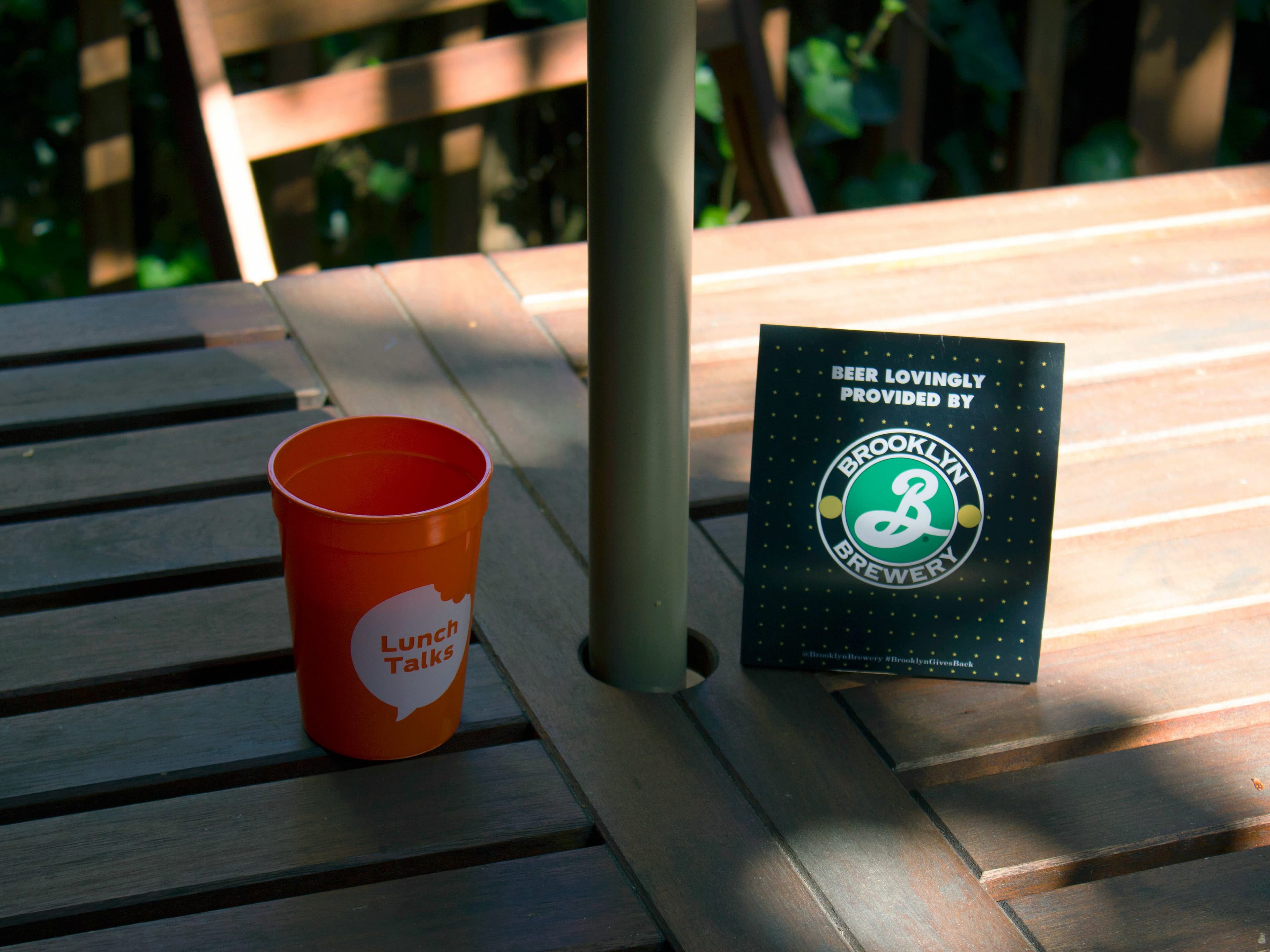 A wooden outdoor table with an orange cup labeled "Lunch Talks" and a sign featuring the Brooklyn Brewery logo. Sunlight and shadows cover the table, creating a calm, inviting atmosphere.