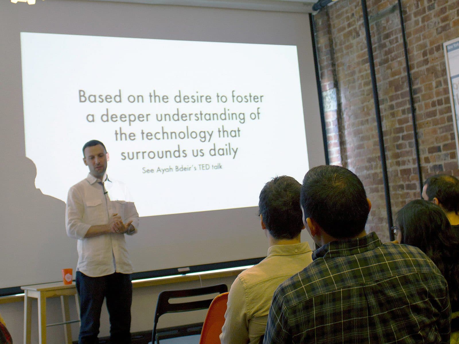 A man in a white shirt stands in front of a screen giving a presentation to an audience. The screen displays the text: "Based on the desire to foster a deeper understanding of the technology that surrounds us daily. See Ayah Bdeir’s TED talk.