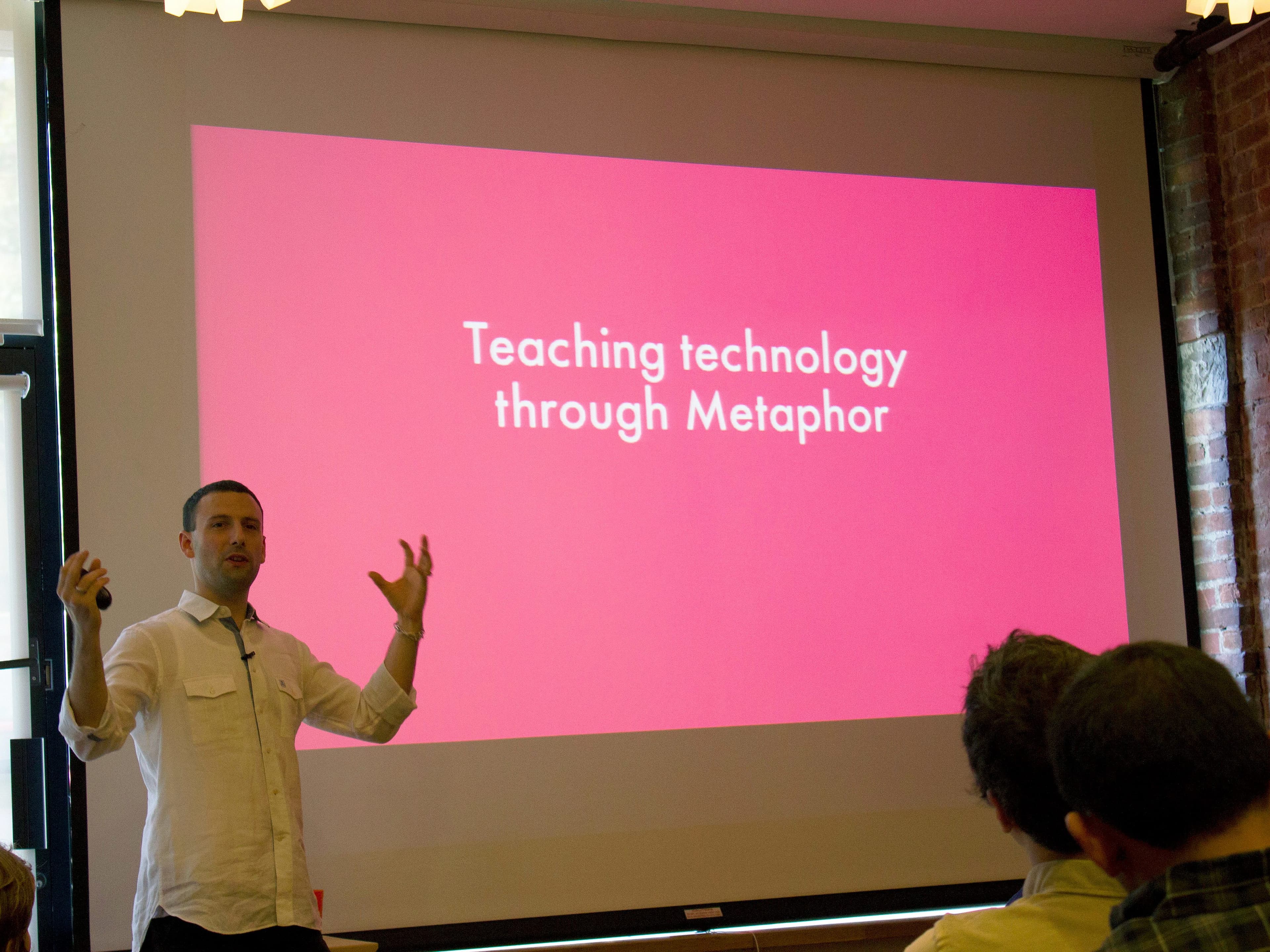 A person in a white shirt is presenting in front of a large screen that displays the slide, "Teaching technology through Metaphor." The screen is pink with white text. The presenter is using hand gestures, and the audience is seated, attentively watching.