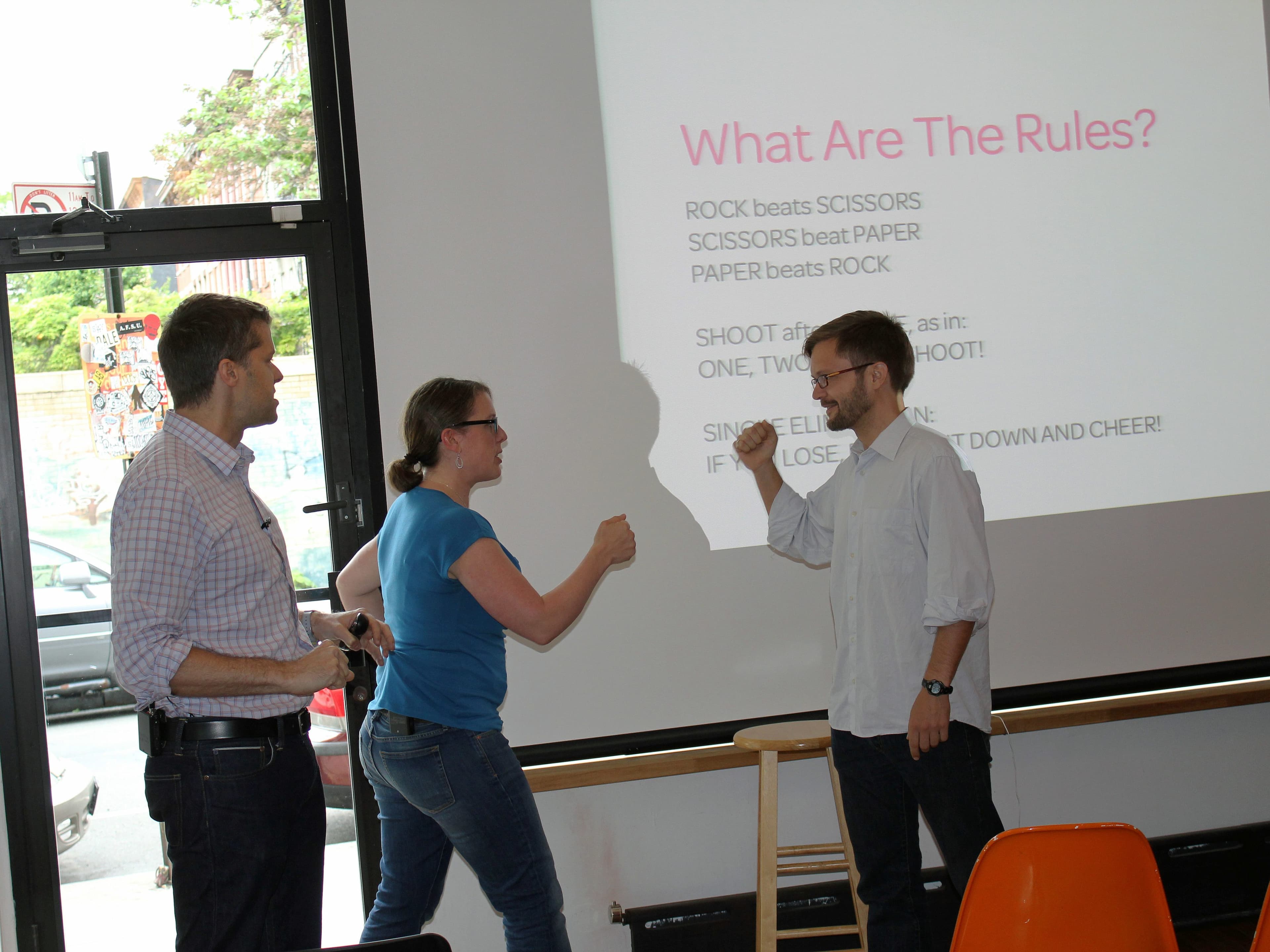 Three individuals are engaged in a rock-paper-scissors game in front of a projected screen displaying the rules. One person is holding a microphone, while the other two are poised to play. Orange chairs are visible in the foreground.