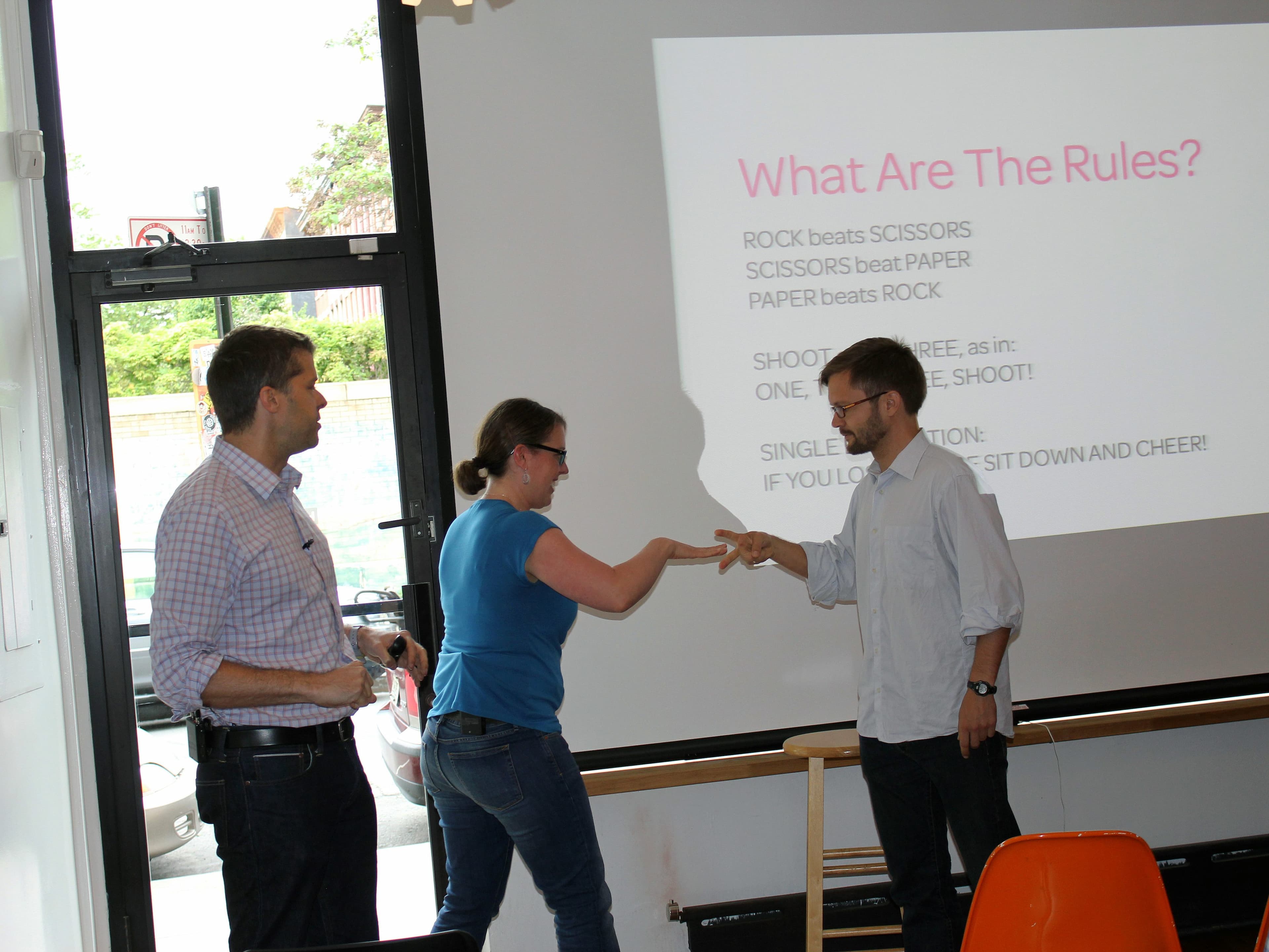 Three people are playing Rock-Paper-Scissors inside a room. A woman in a blue shirt and a man in a white shirt are making hand gestures, while another man in a checked shirt watches. Behind them, a screen displays the rules of the game.