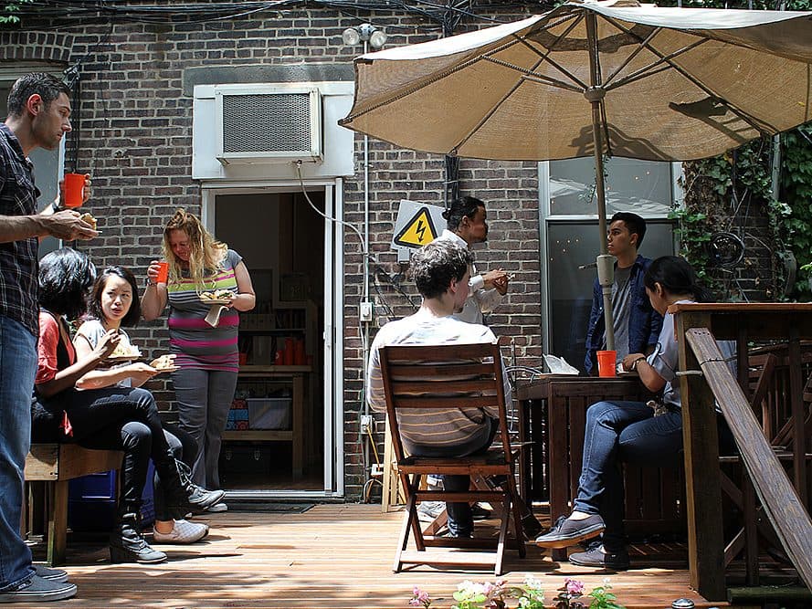 A group of people are enjoying food and conversation on a sunny patio. Some are sitting at a table under a large umbrella, while others stand or sit nearby. The background features a brick building with an open door and windows. 

.